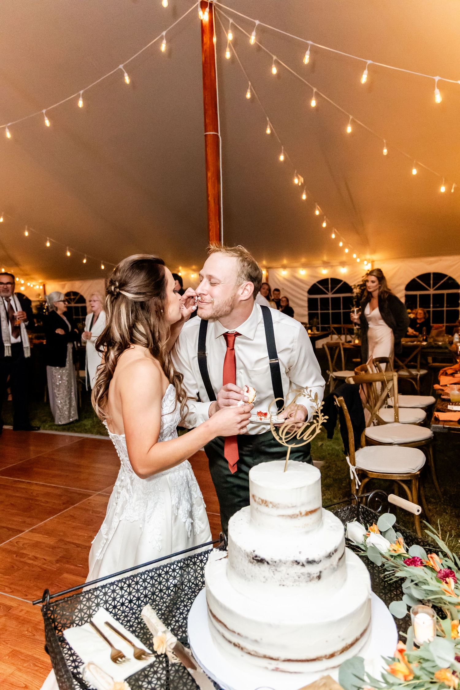 Couple shares a playful moment while cutting their wedding cake under twinkling string lights at an outdoor reception.
