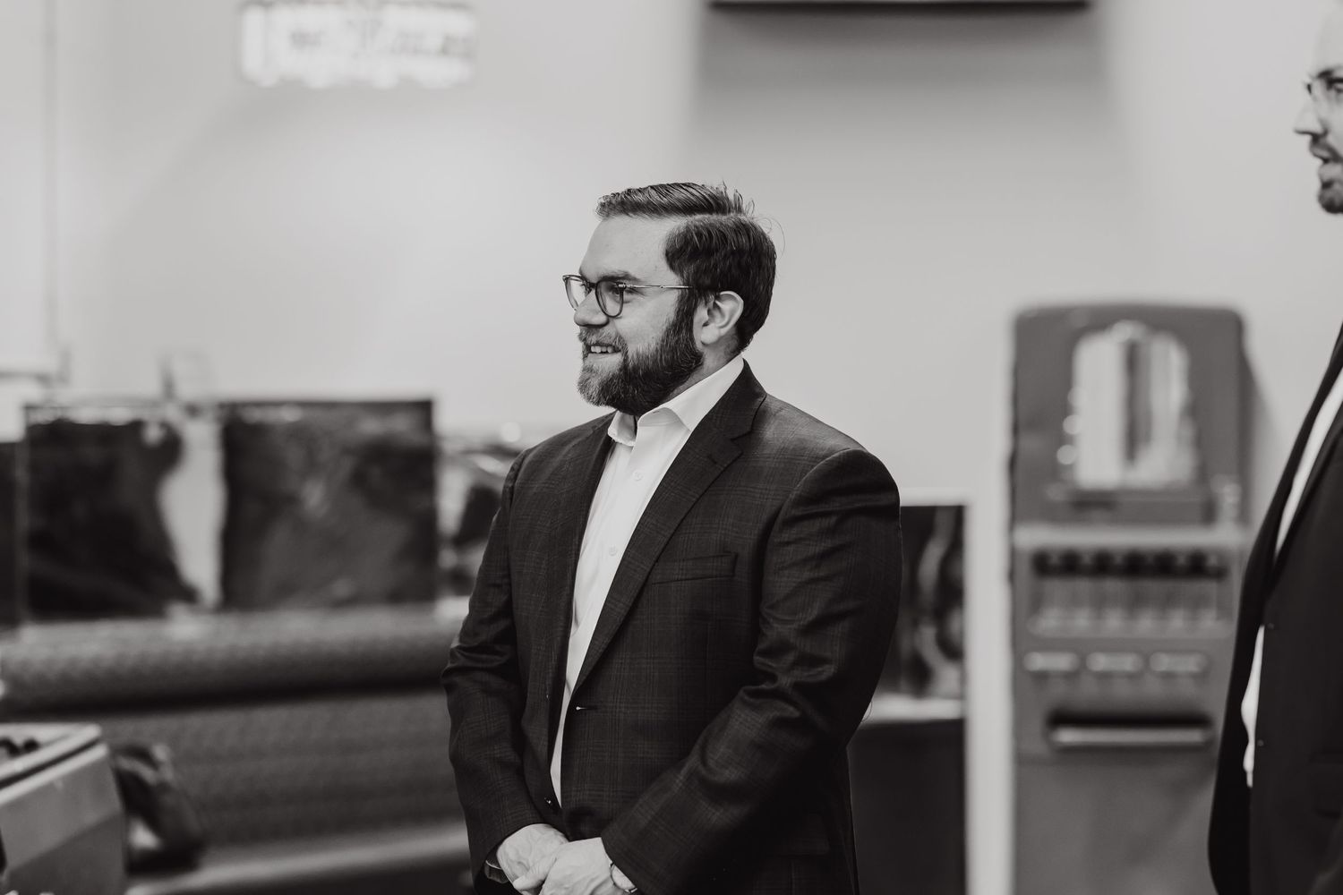 A man in a dark suit and glasses stands in a dimly lit indoor setting.