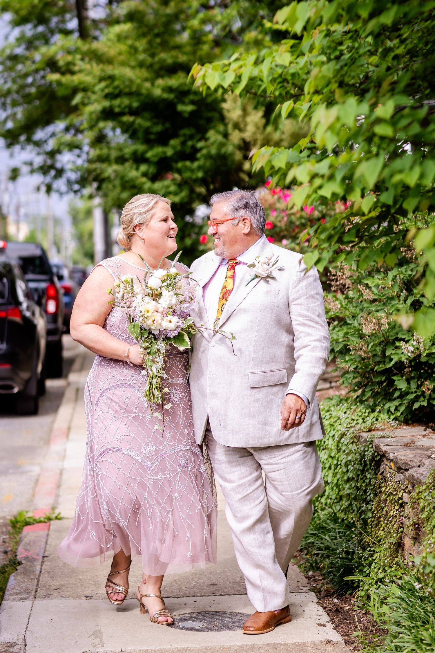 A couple in formal attire walking together on a sidewalk, with the person in rose pink holding a bouquet.