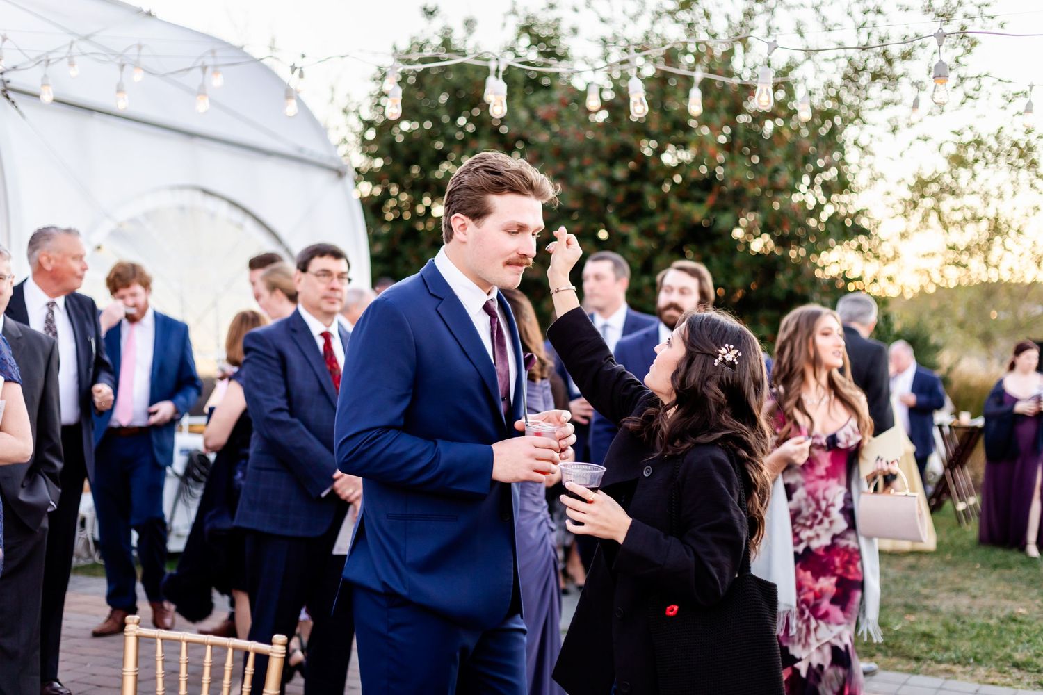 Wedding guests mingle during outdoor cocktail hour near white tent at sunset.