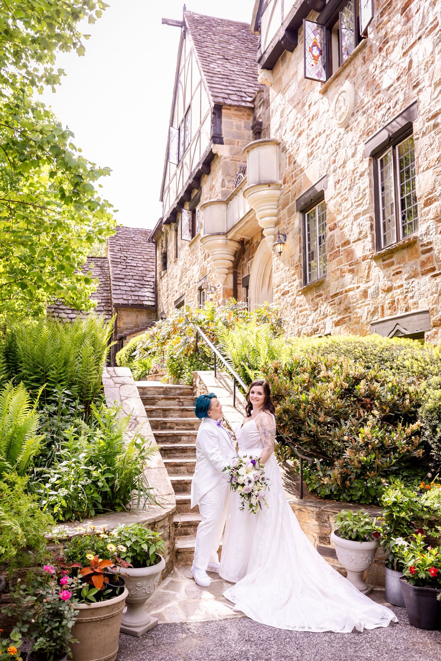 Newlyweds pose on garden steps of a historic stone building with potted flowers and greenery.