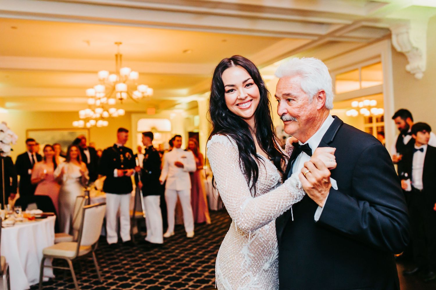 Two people share a heartfelt dance moment at an elegant indoor reception with guests looking on in the background.