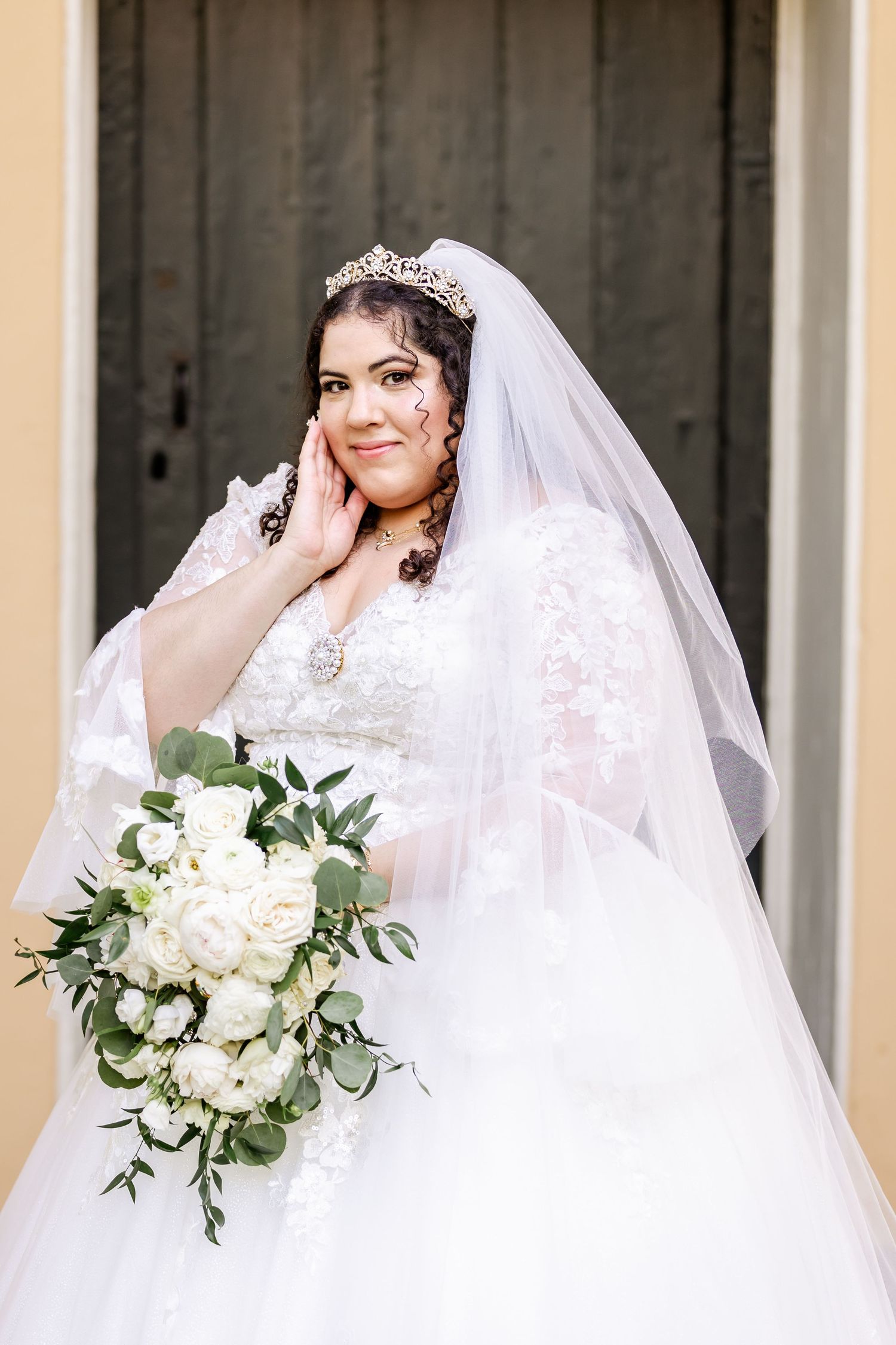 A bridal portrait showcasing an elegant lace wedding dress, delicate tiara, and white floral bouquet with greenery.