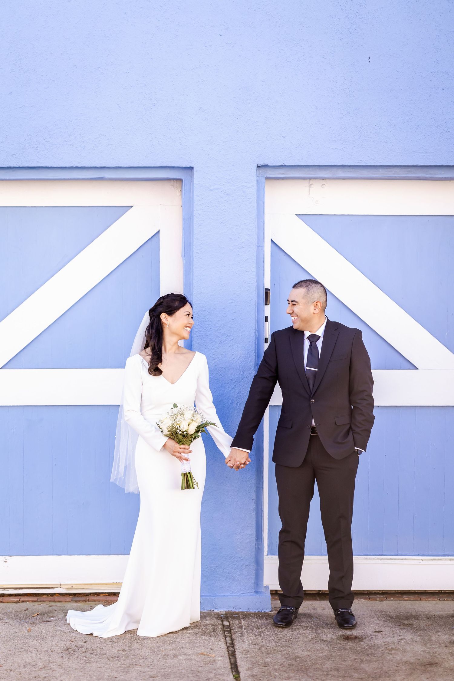 Newlyweds pose against a blue and white geometric wall holding a wedding bouquet.