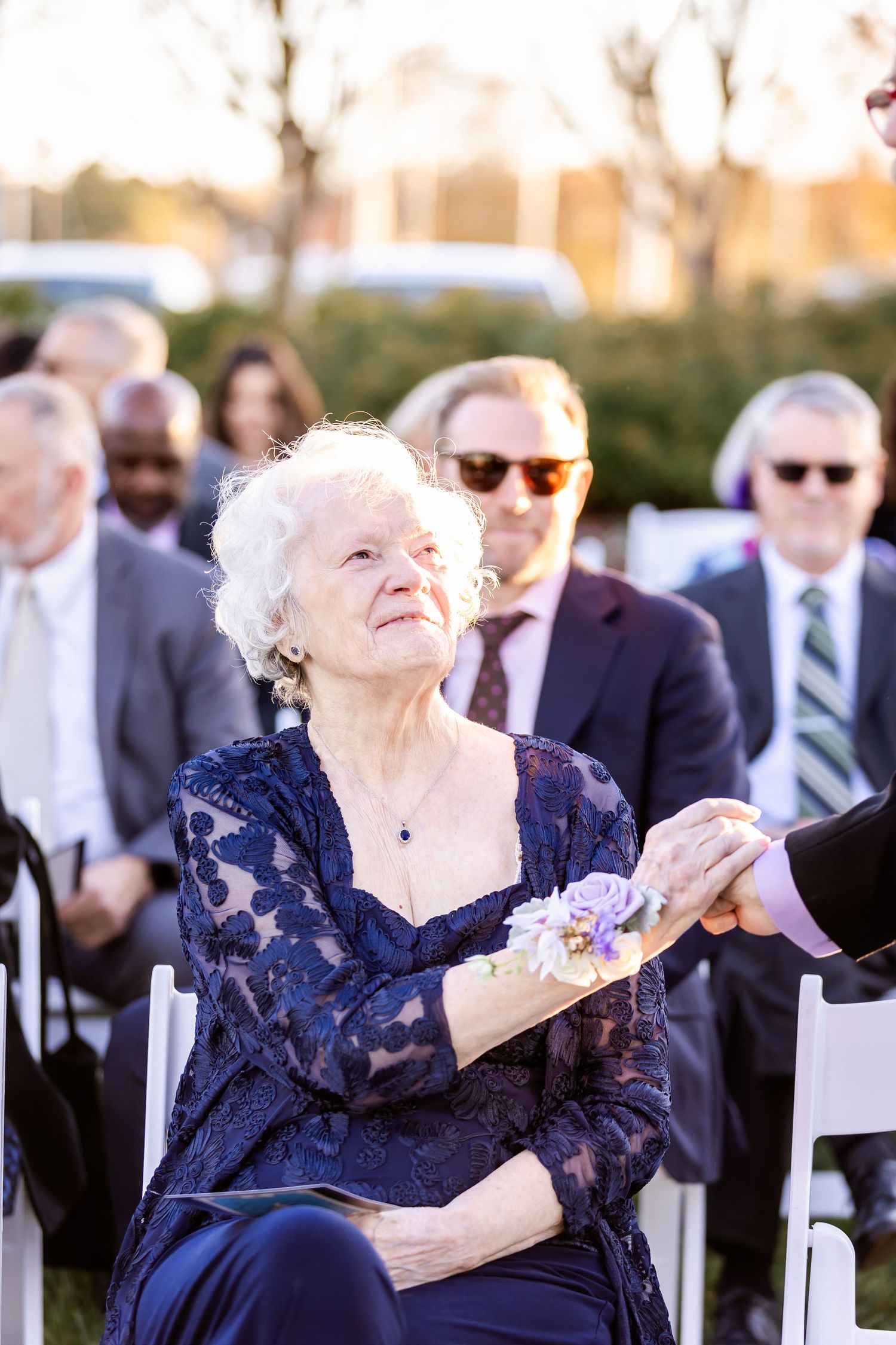 Senior woman in navy lace dress sits elegantly at outdoor ceremony.