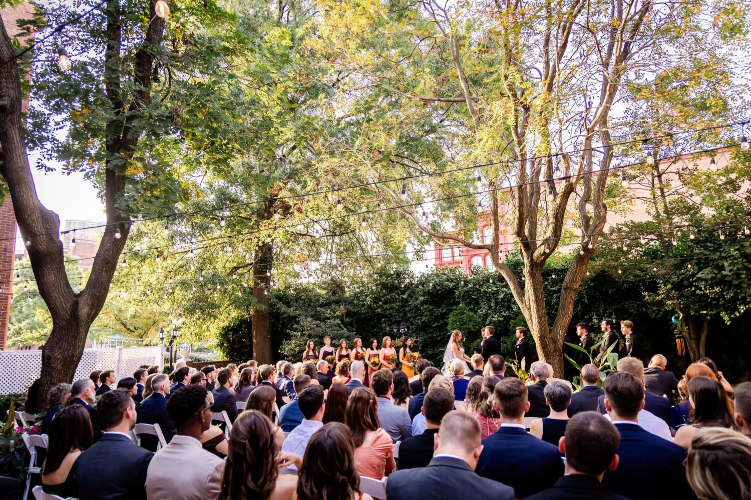 A large crowd gathers outdoors under trees for a wedding ceremony on a sunny autumn day.