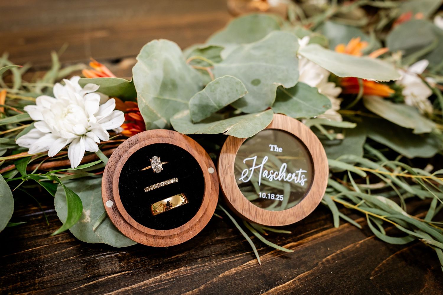 Wedding rings displayed in wooden ring boxes surrounded by eucalyptus leaves and white flowers on dark wood surface.