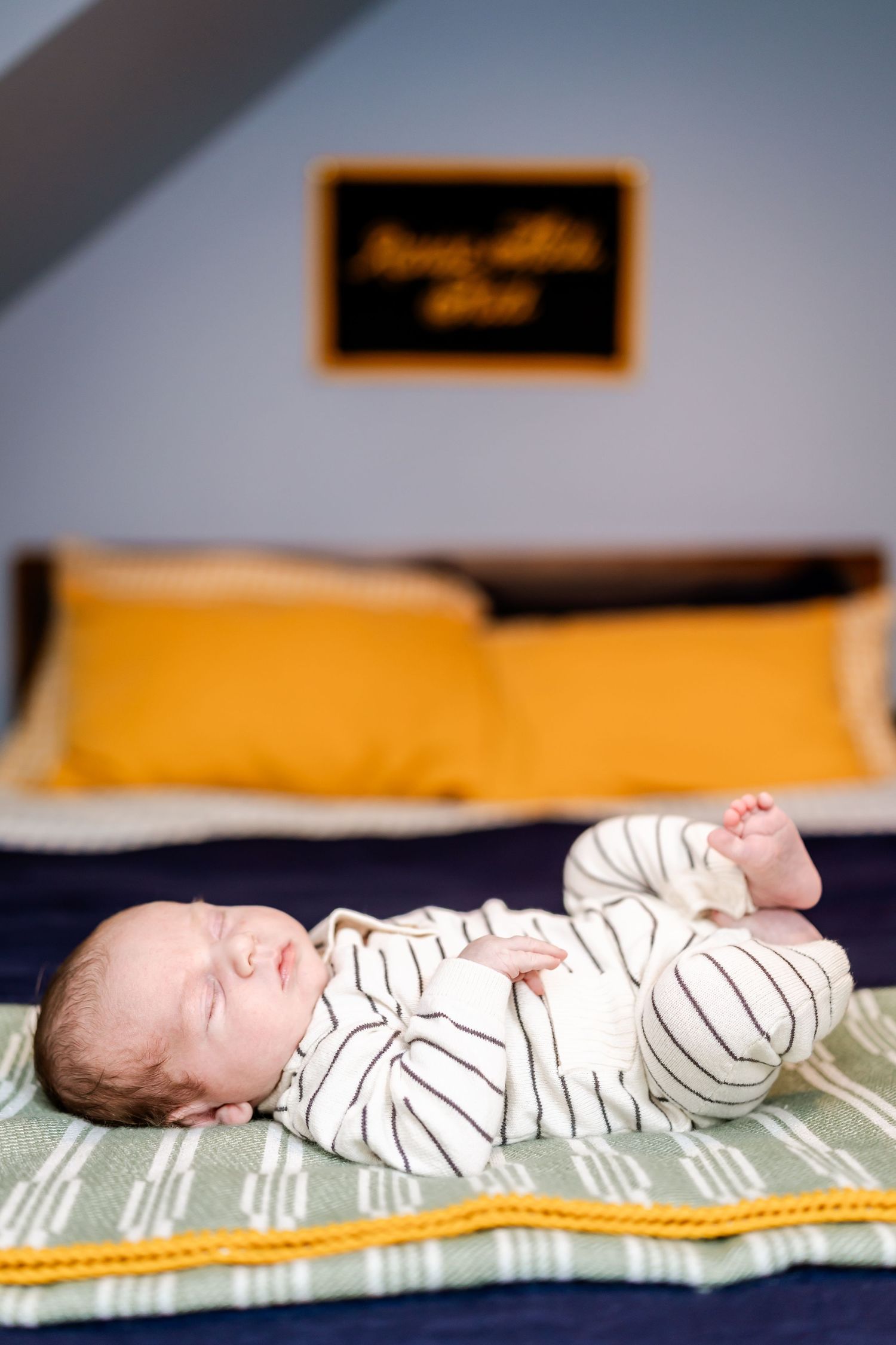 Sleeping newborn baby in striped onesie lying on blue bedding with yellow pillows in background.