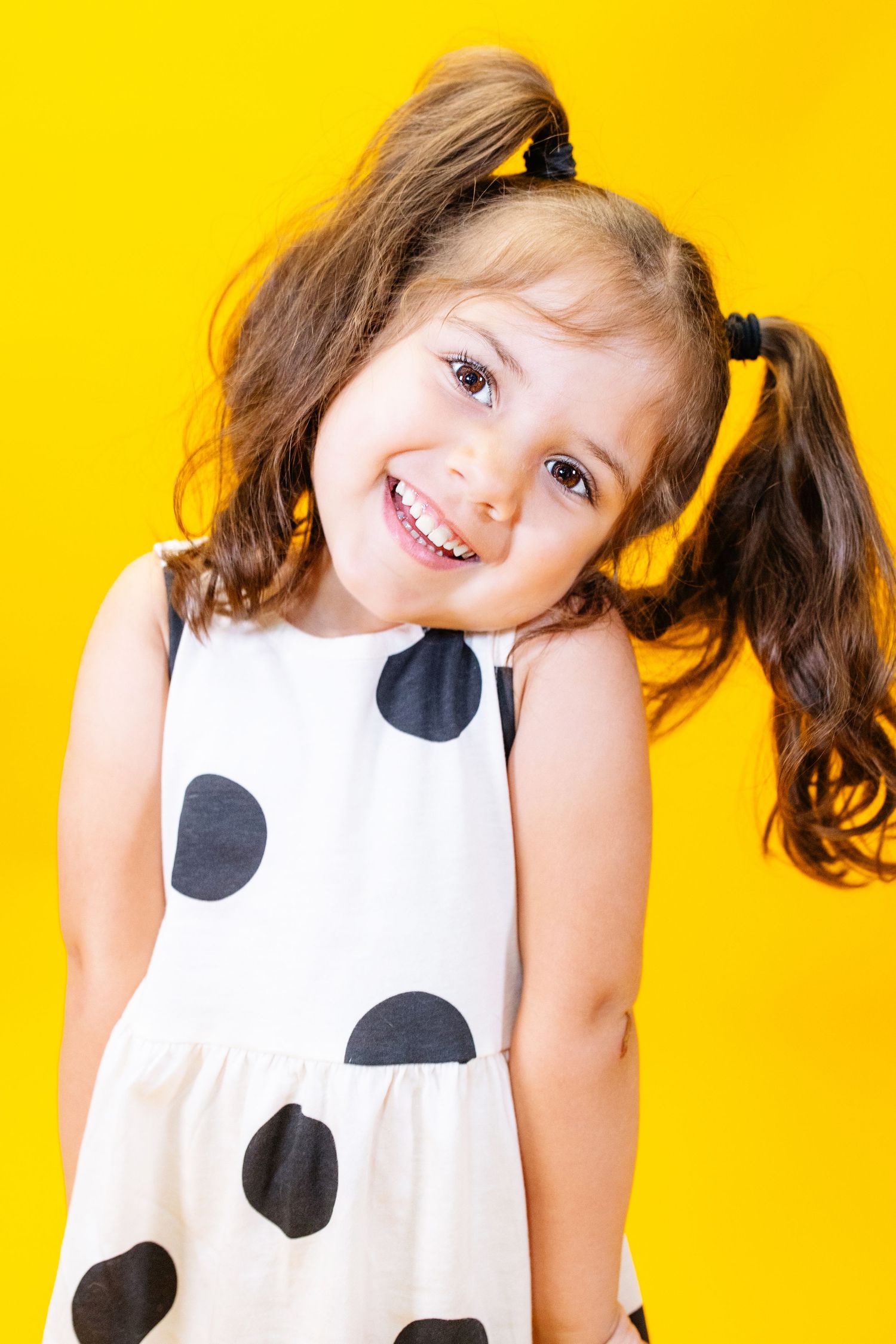 A child makes silly expressions while wearing a black and white polka dot dress against yellow backdrop.