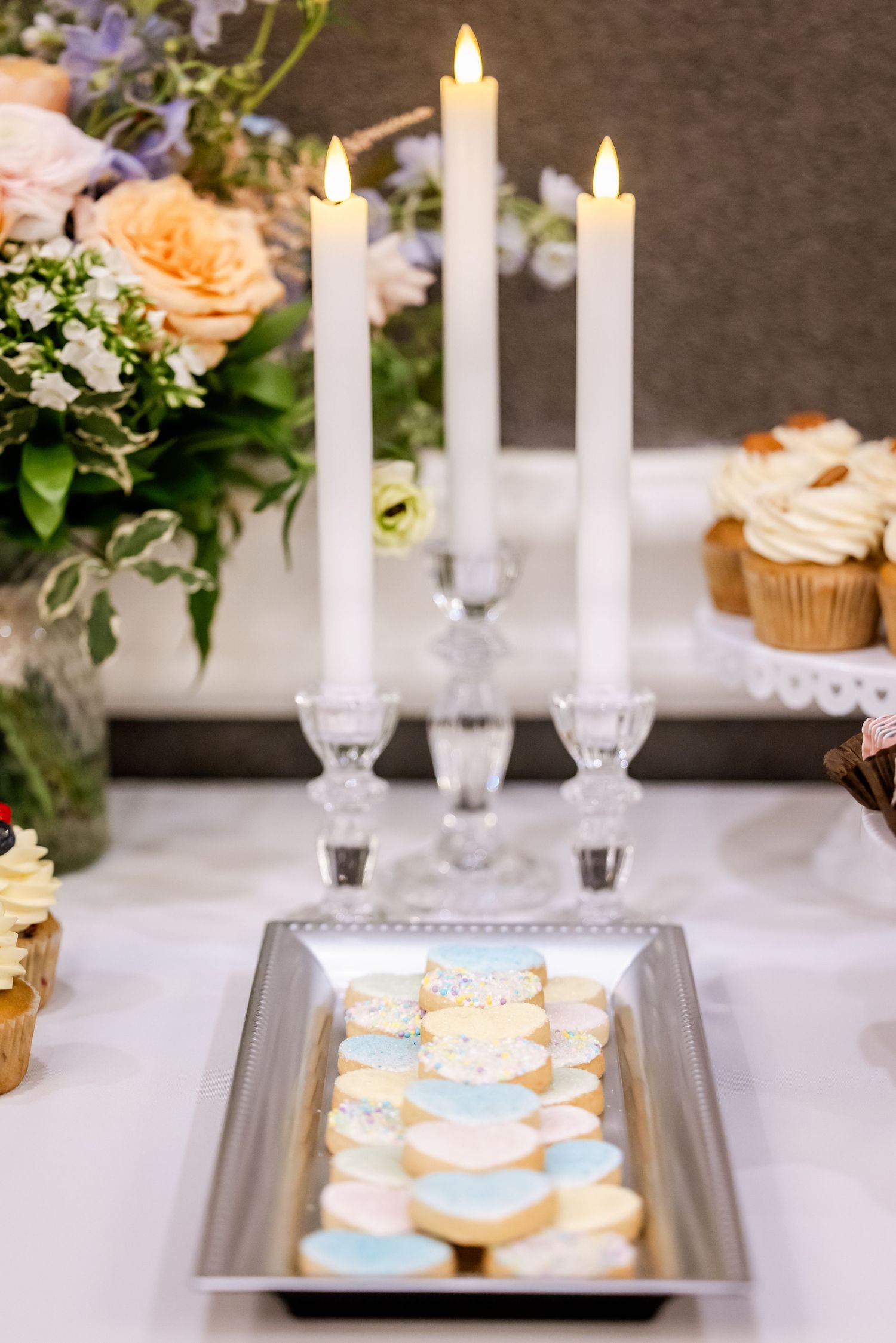 Elegant wedding reception table with white taper candles, floral arrangements, and decorated cookies on silver platters.