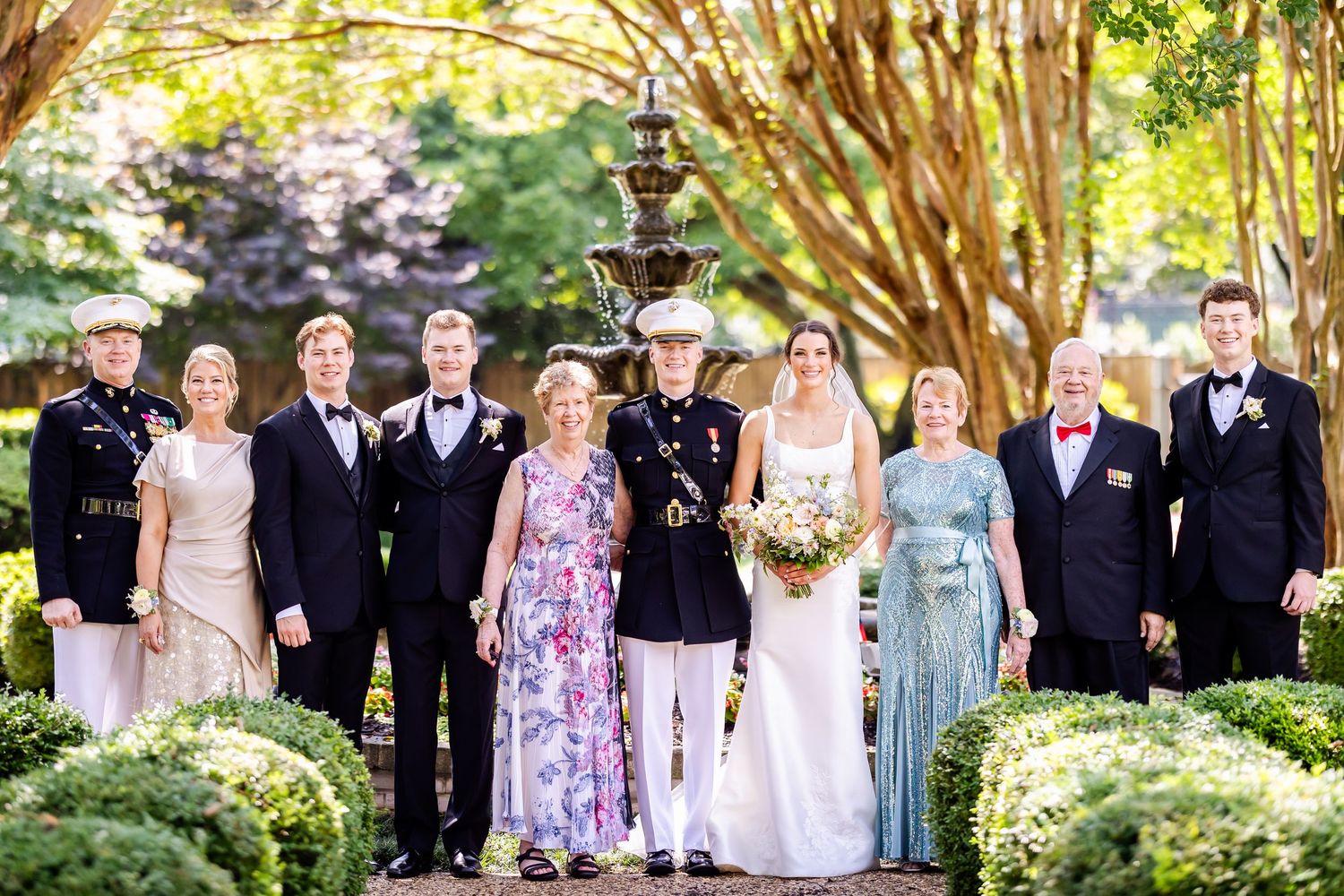 Wedding party poses together outdoors in formal attire with military dress uniform against lush garden backdrop.