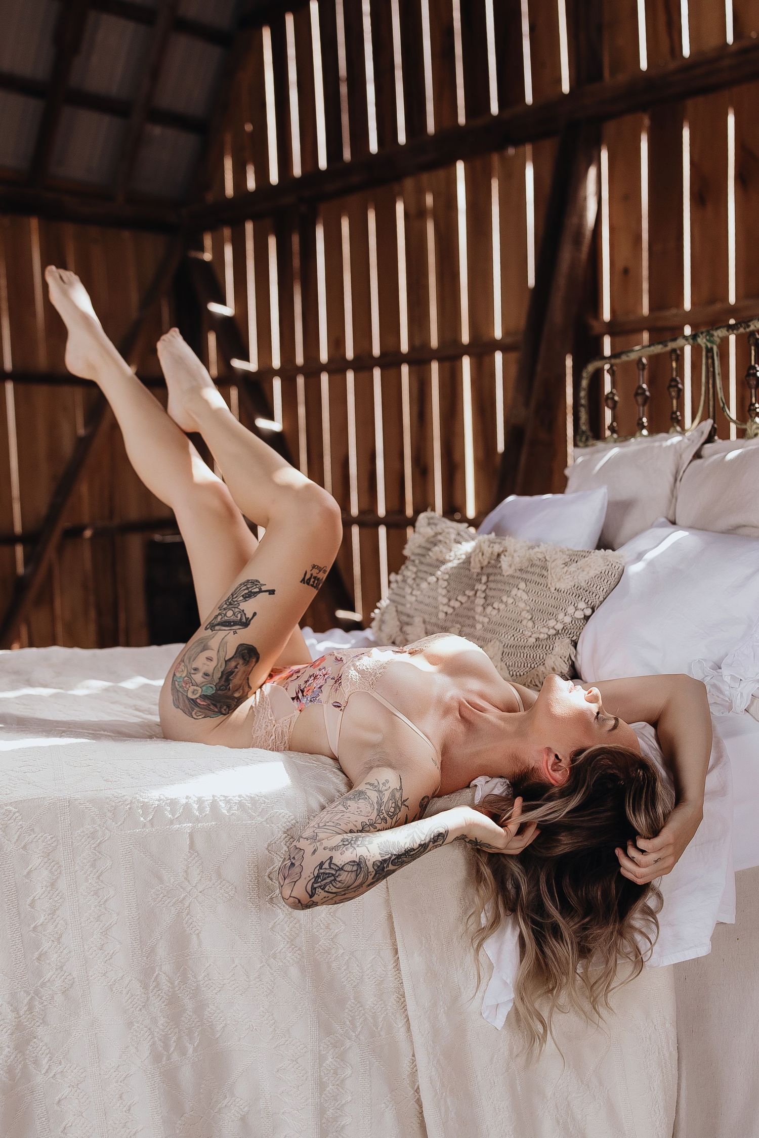 Woman with tattoos in lingerie posing on white bed in rustic wooden barn setting with natural light.