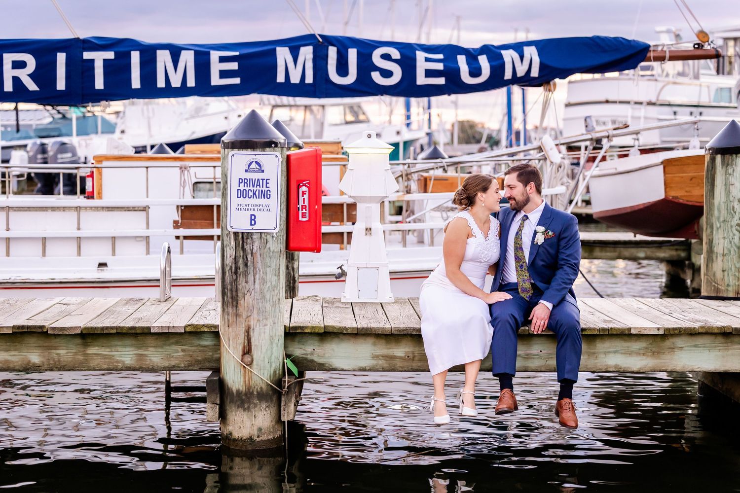 Bride and groom sitting on a dock edge at the Maritime Museum, smiling.
