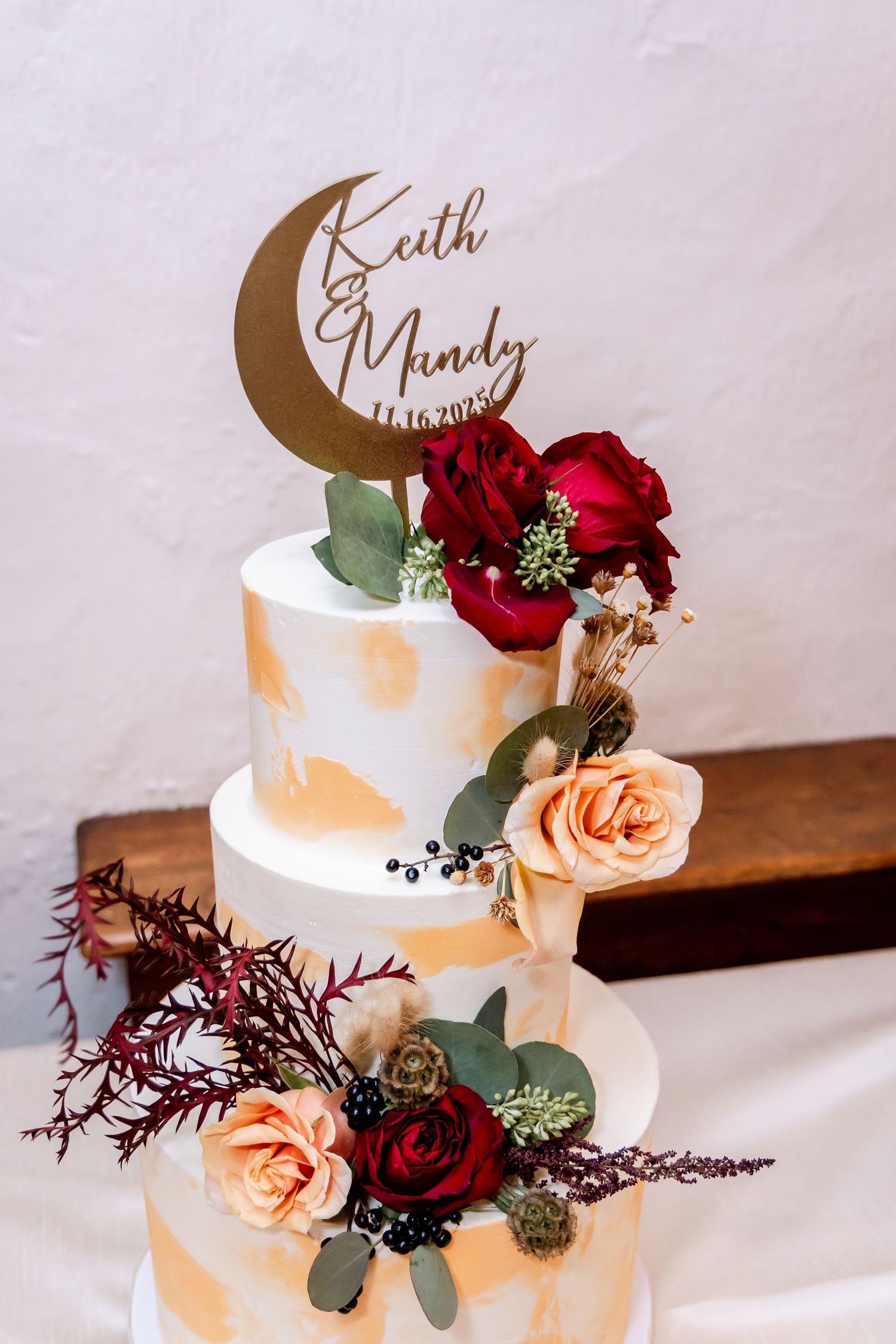 Three-tier white wedding cake decorated with burgundy and peach roses and gold crescent moon topper.