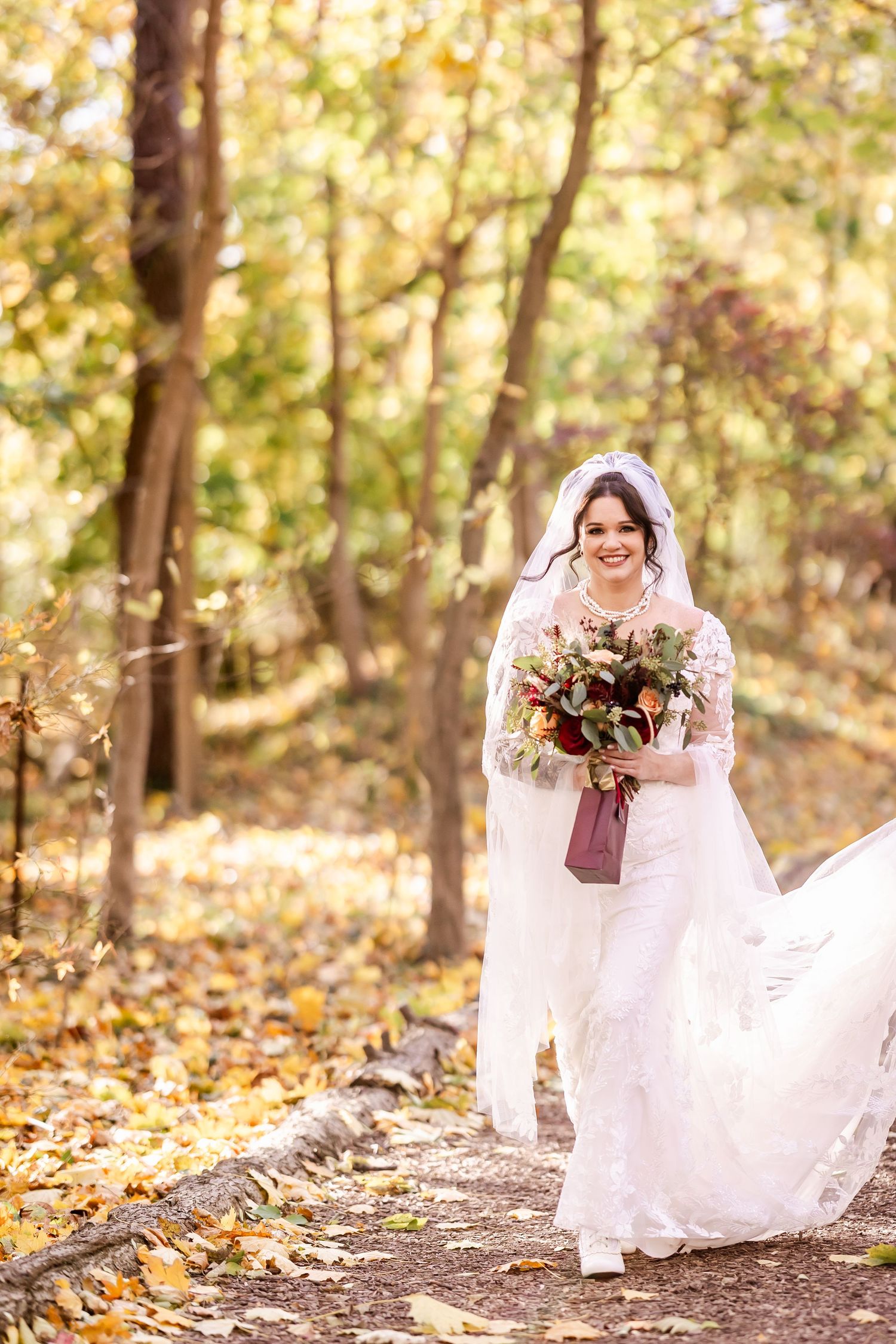 A bride in a flowing white dress holds an autumn bouquet while walking along a wooded path covered in fallen leaves.