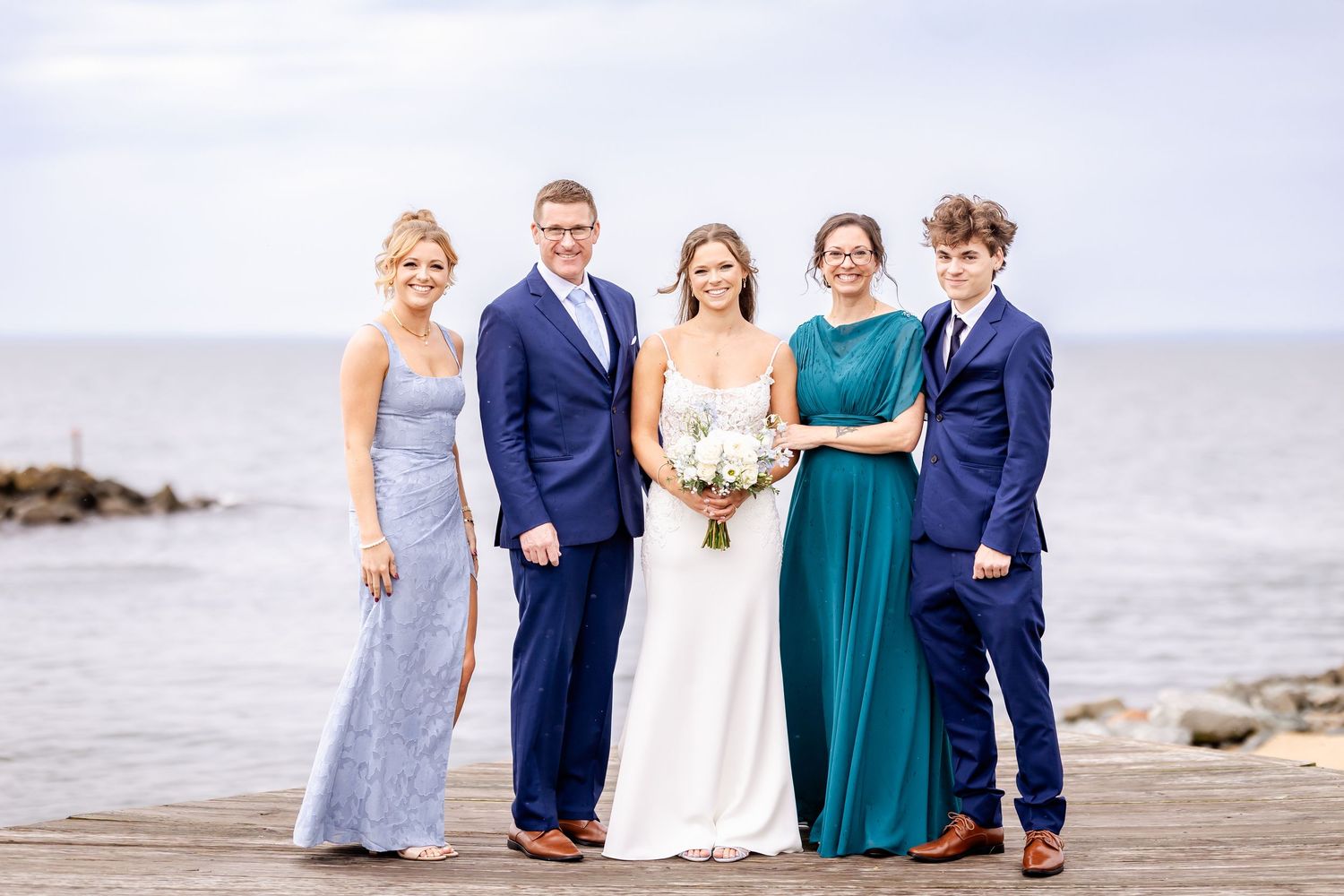 A wedding party poses together on a rocky beach with ocean waves and cloudy skies in the background.