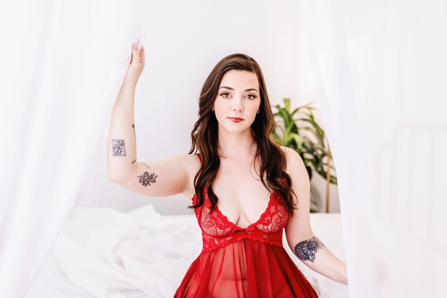 Woman in red lingerie poses elegantly on white bedding in bright boudoir setting.