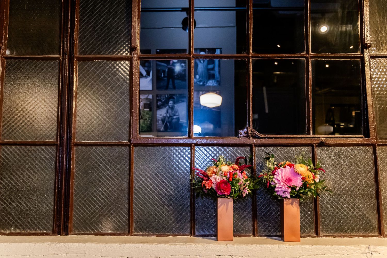 Two floral arrangements in wooden vases sit beneath industrial windows with wire mesh at night.