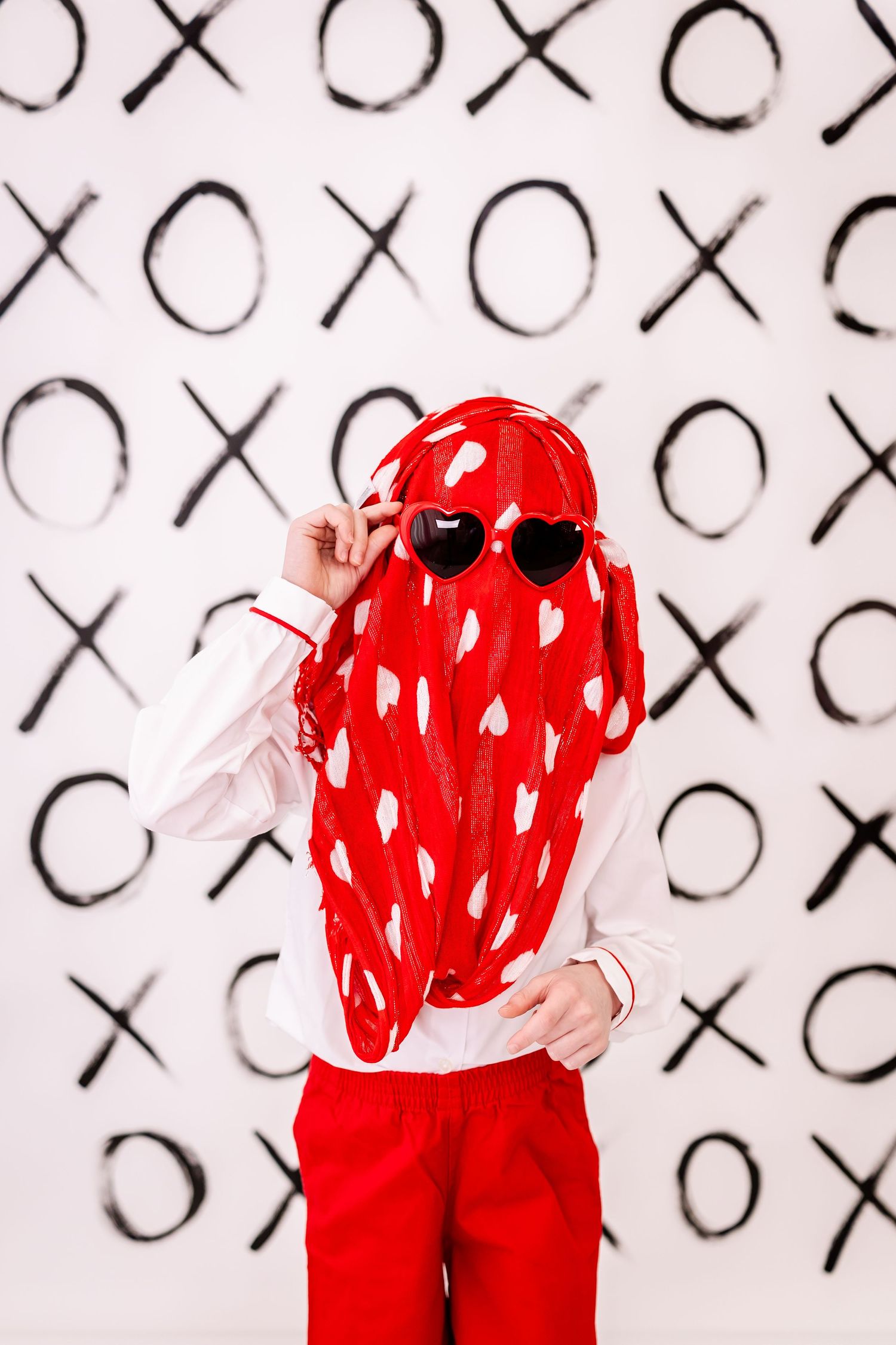 Artistic portrait against XO pattern backdrop wearing red and white heart-patterned outfit and red sunglasses.