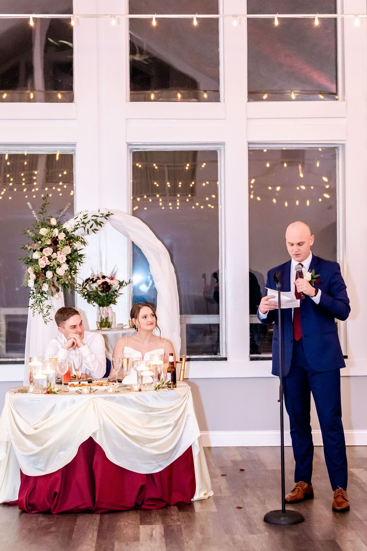 Wedding reception setup with elegant white draping and burgundy accents in bright venue.