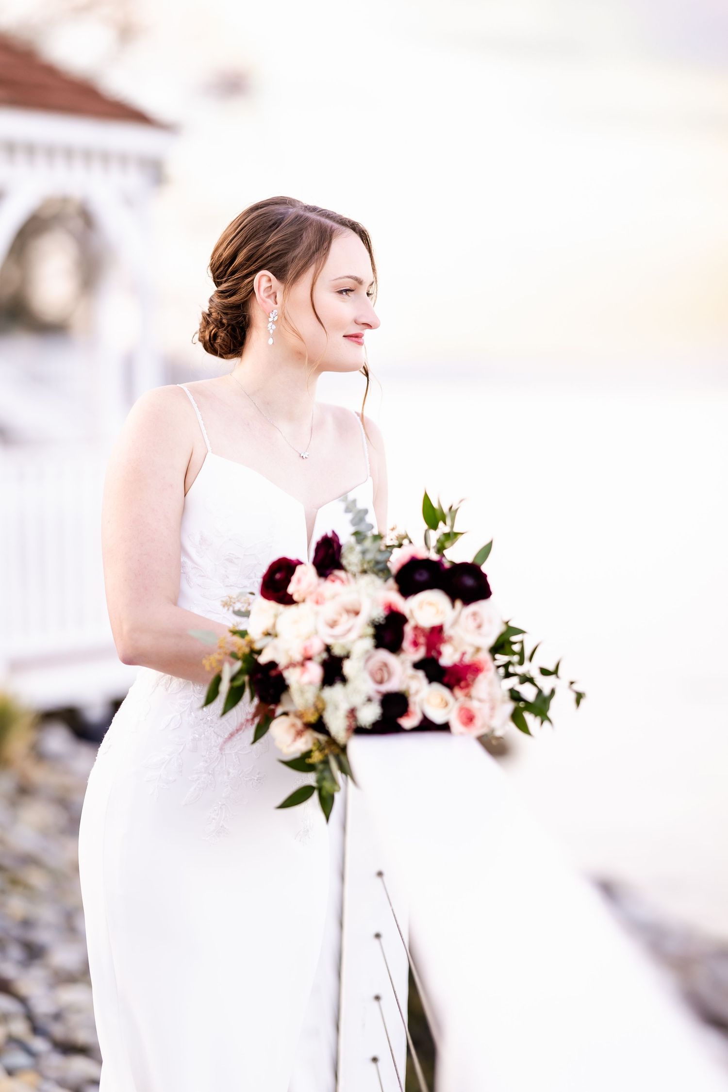 Elegant bride in white dress holds romantic bouquet with burgundy and blush flowers against light background.