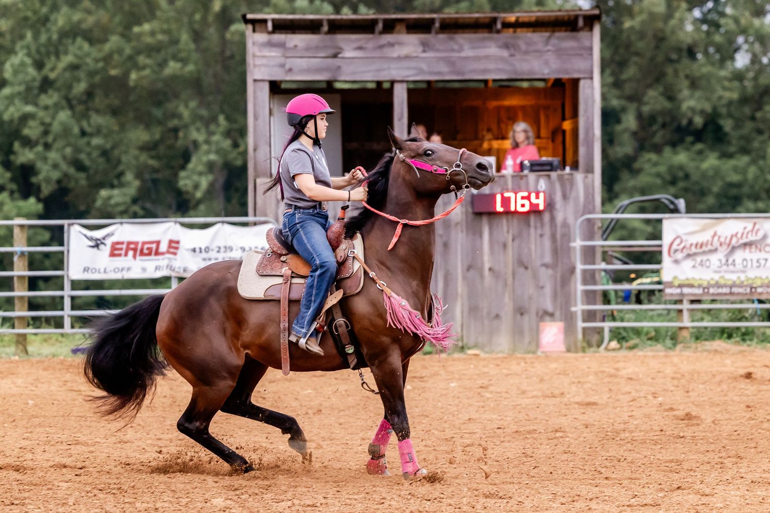 Horse and rider perform barrel racing maneuvers in a dusty outdoor arena near rustic structure.