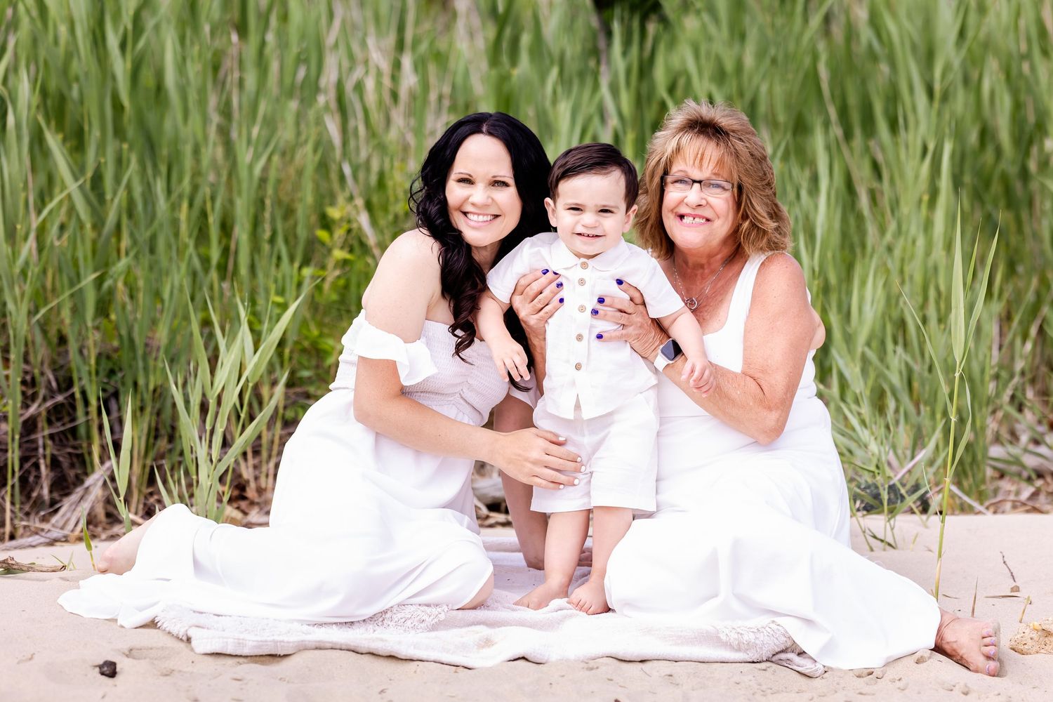 Three people in white dresses pose together on a sandy beach with tall grass in the background.
