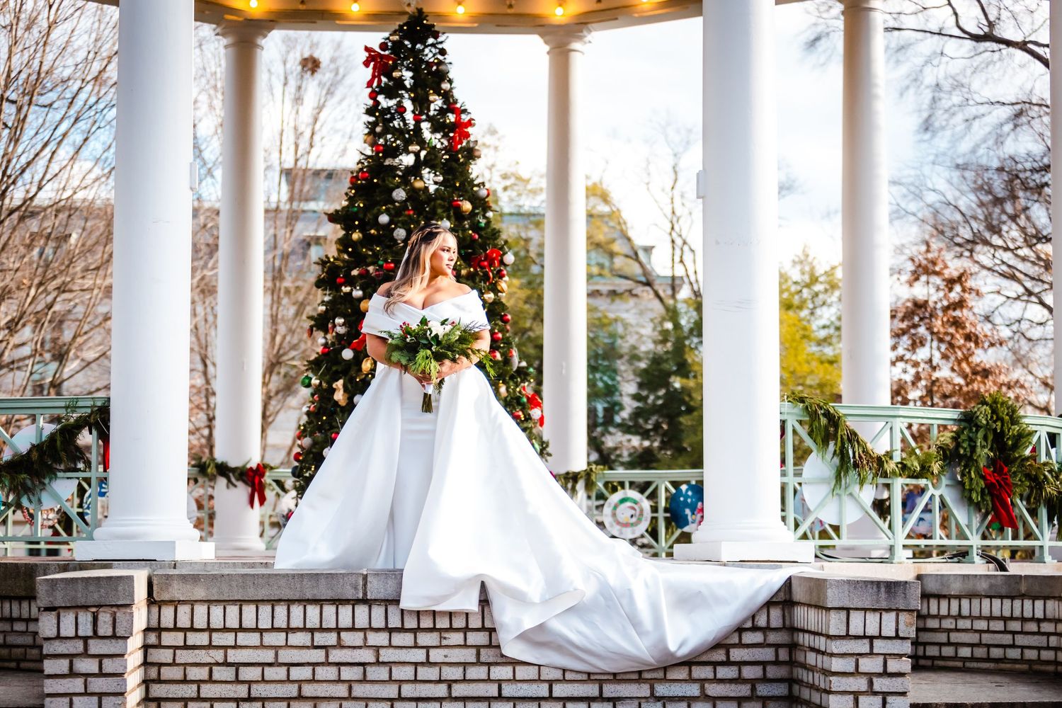 A white wedding gown with long train in front of a decorated Christmas tree on a brick gazebo with white columns.