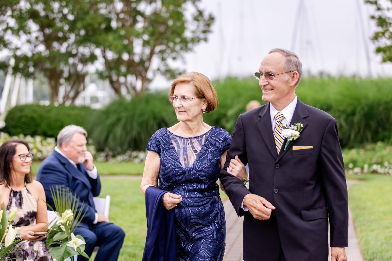 Wedding guests in navy blue and dark attire gather outdoors on a sunny day during a celebration.
