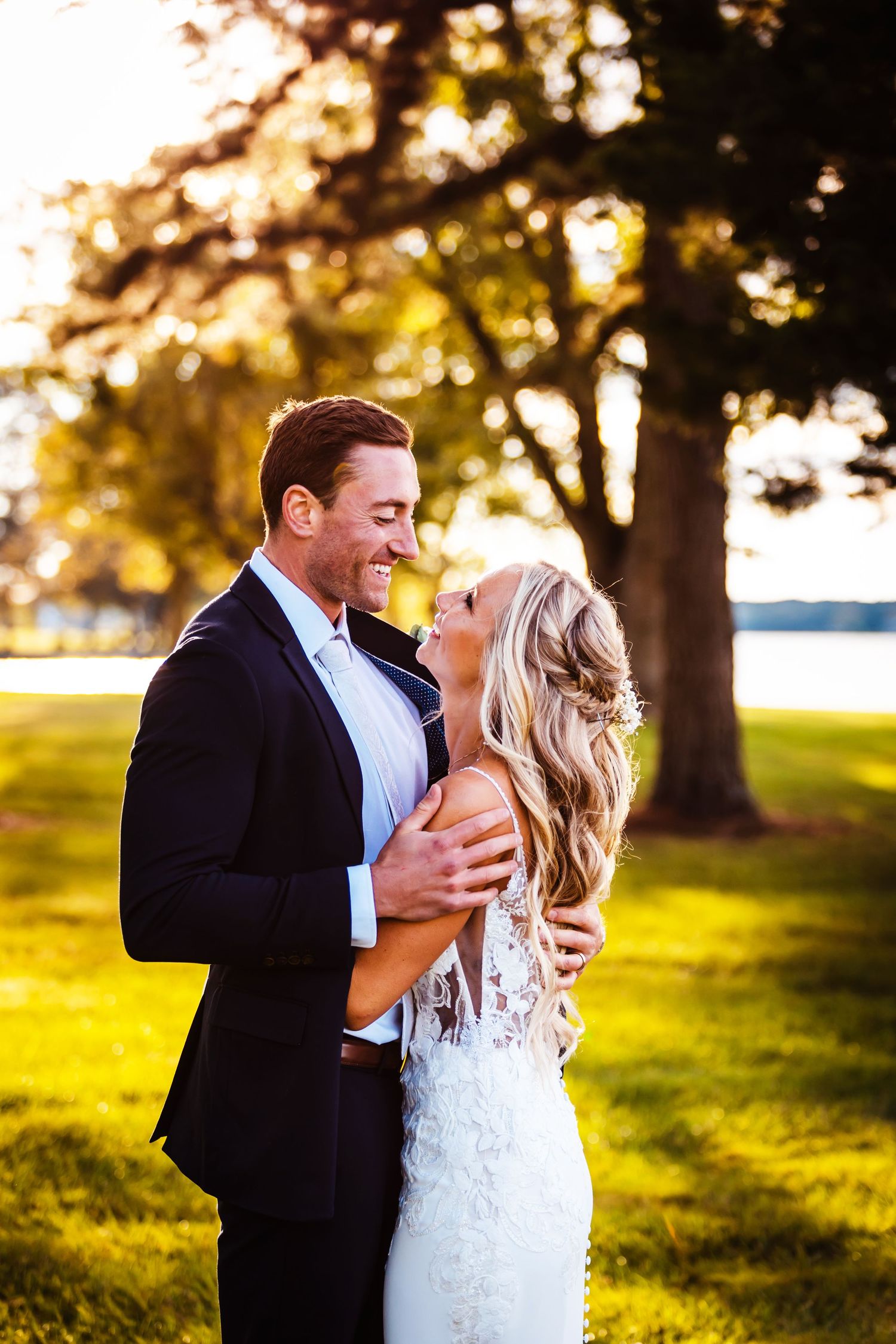 A couple shares a joyful moment in formal wedding attire at sunset with trees and a lake in the background.