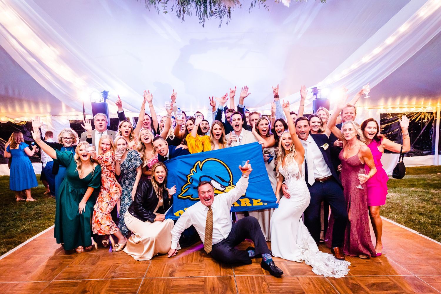 Large wedding party celebrating and posing together on a dance floor under an elegant white tent with ambient lighting.