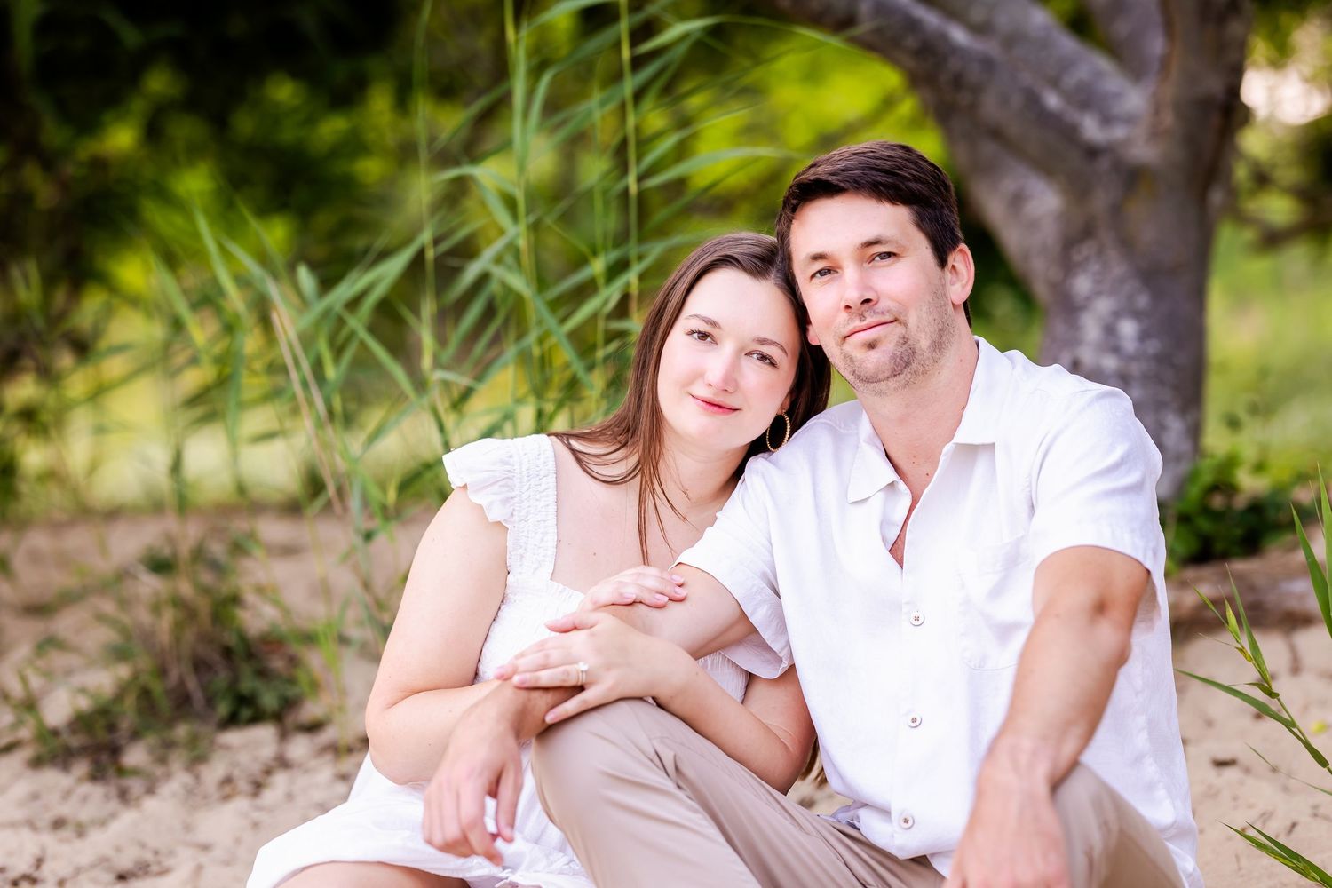 Happy romantic couple in white outfits sitting together outdoors in front of tall grass and trees.