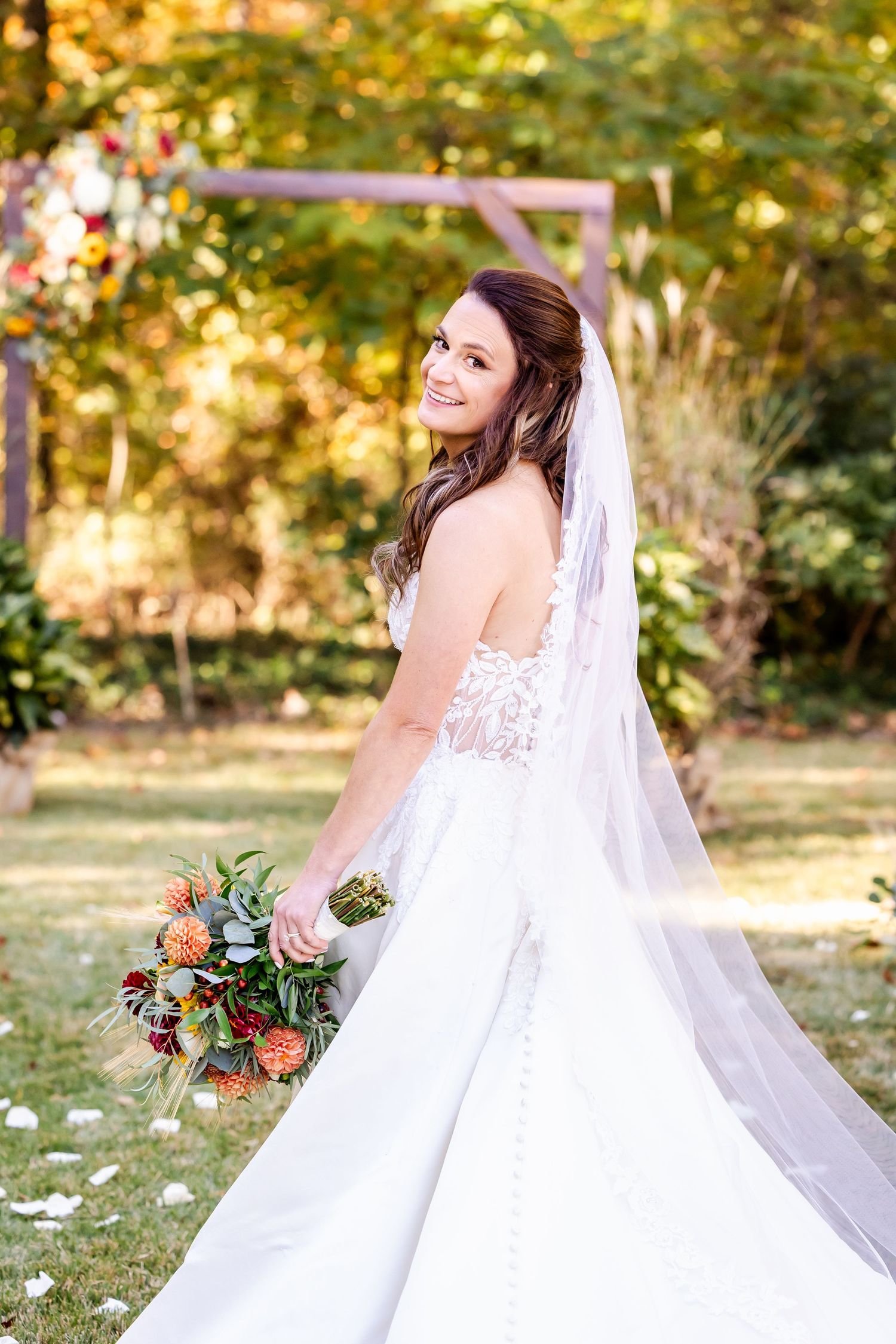 A bride in a white lace gown poses gracefully in an autumn garden setting with golden foliage in the background.