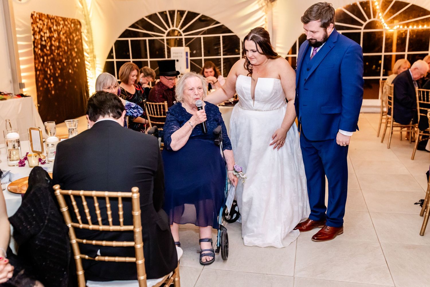 Wedding reception scene showing couple greeting guests in an elegantly decorated venue with string lights and arched windows.