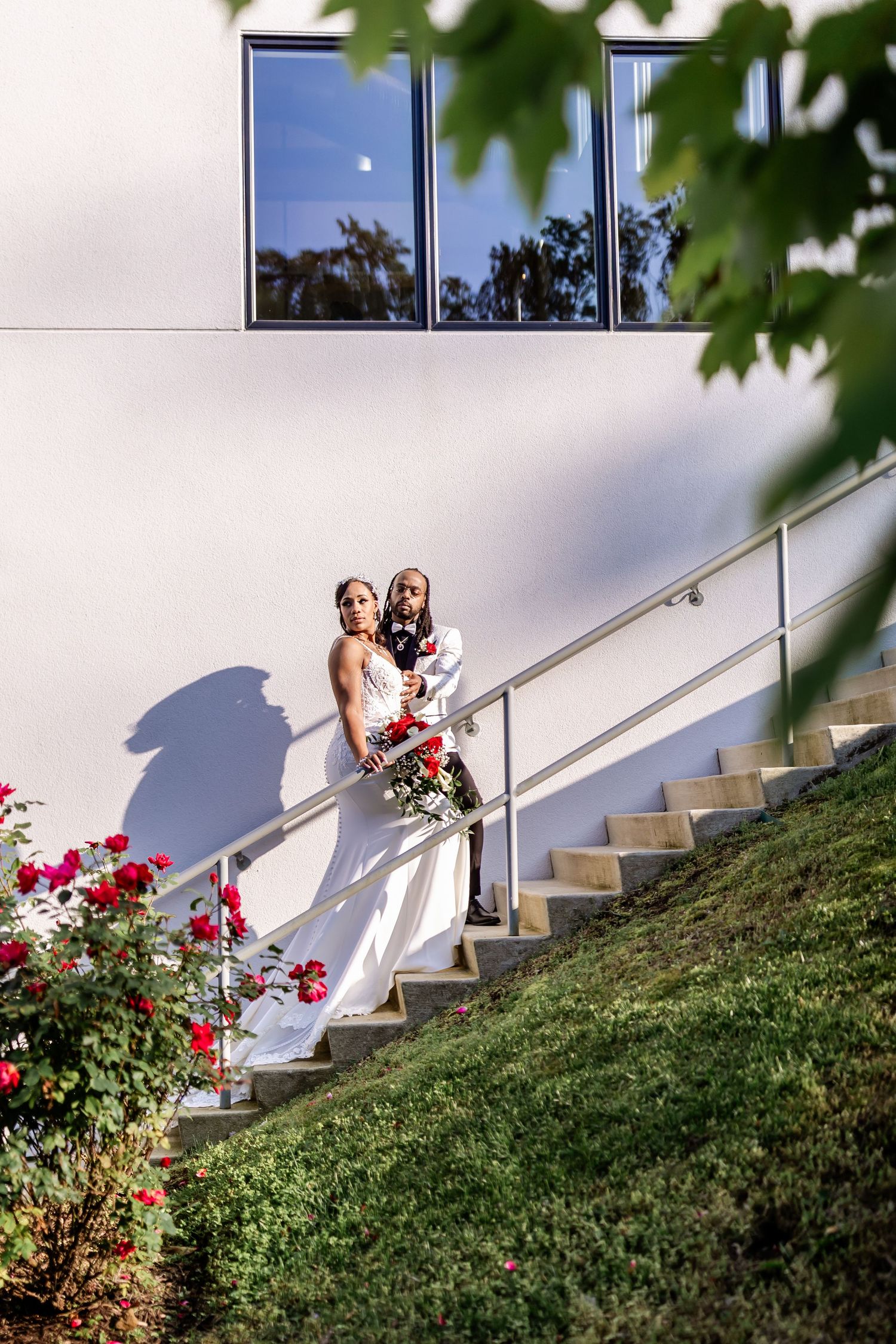 Wedding couple posing on outdoor staircase with red roses and white building backdrop.