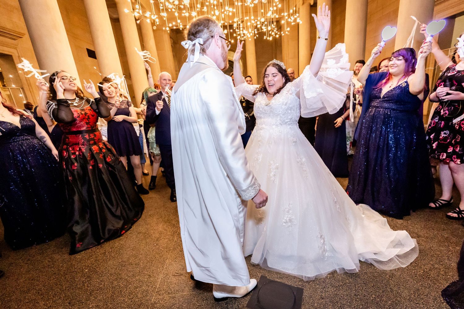 Wedding celebration with guests dancing joyfully around the couple in an ornate hall.