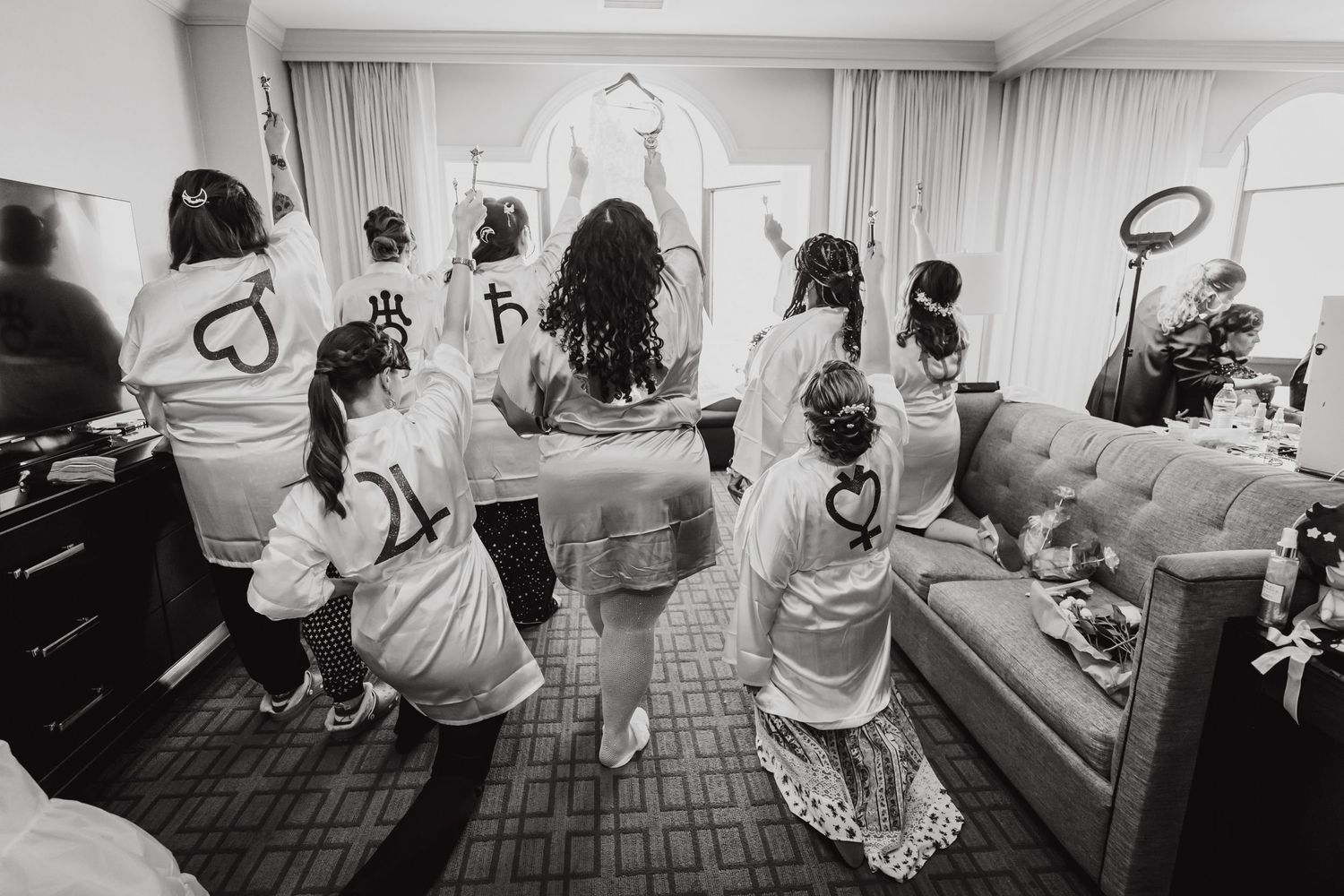 Black and white photo of wedding party dancing in white robes in hotel room.
