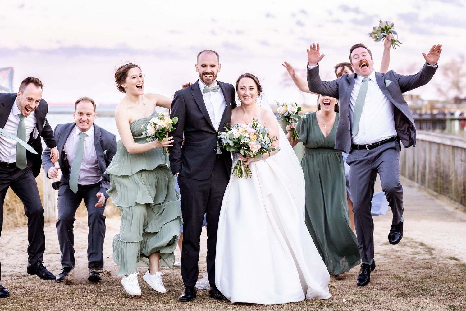A wedding party in green dresses and formal attire jumps and celebrates joyfully together on a beachside path.