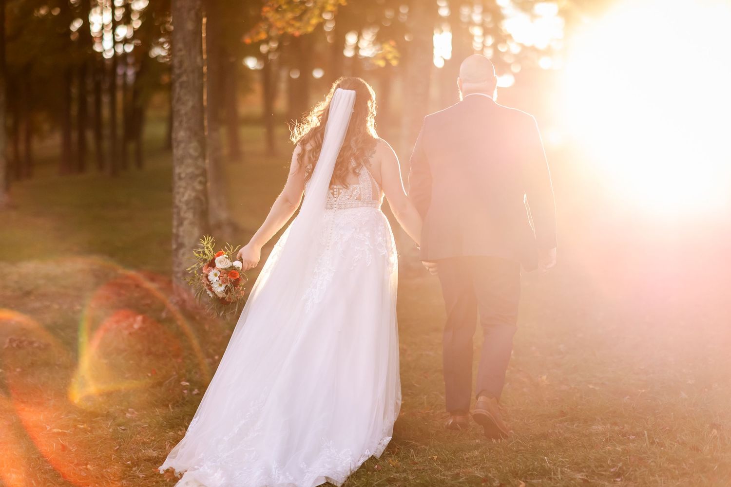 Newlywed couple holding hands and walking through sunlit forest with ethereal golden sunset glow.