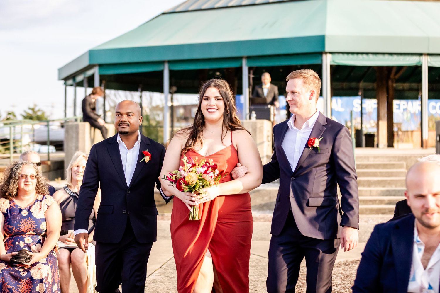 Wedding party processional walking along waterfront path on sunny day.
