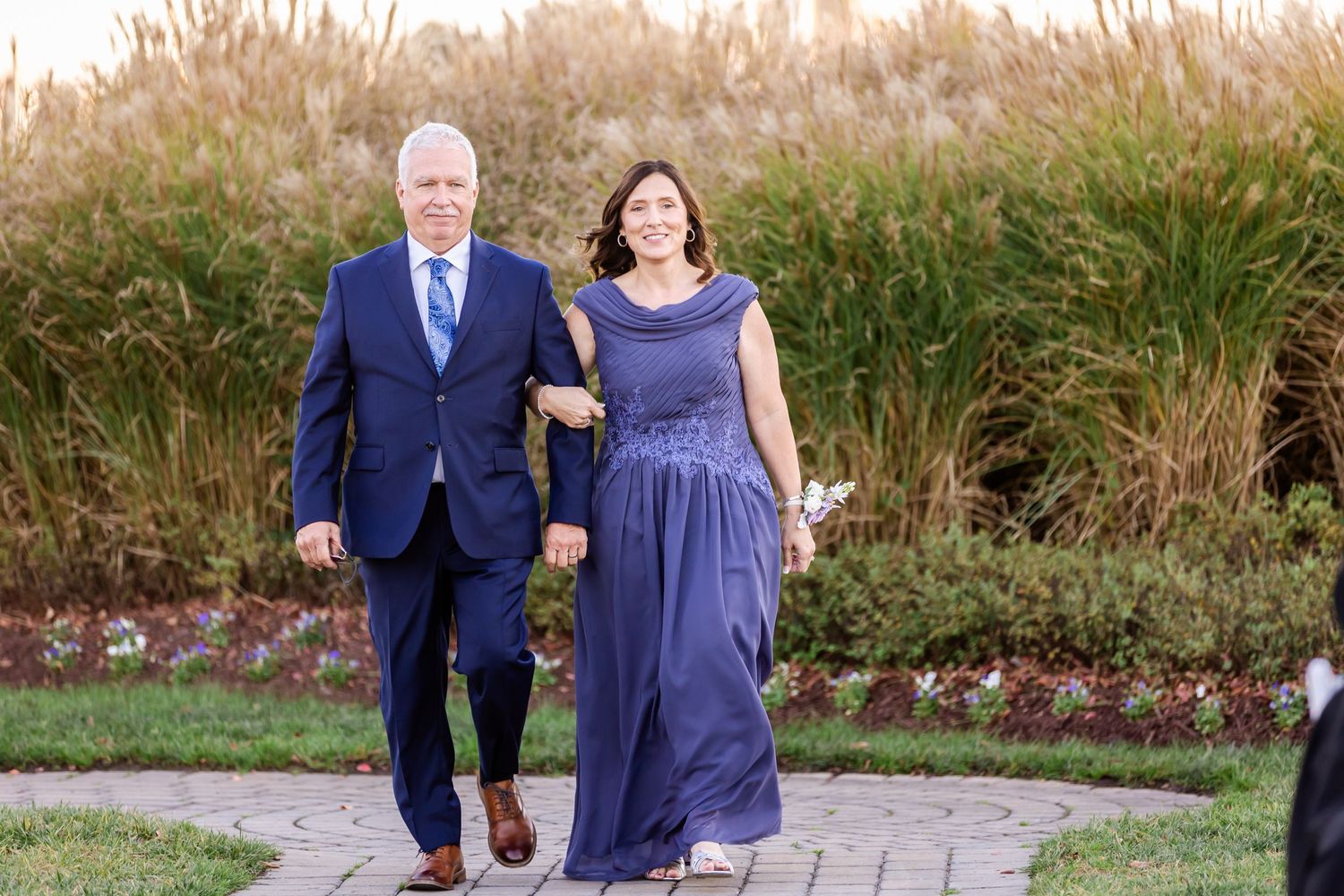 A couple walks arm in arm on a garden path at sunset wearing coordinating formal navy blue attire.