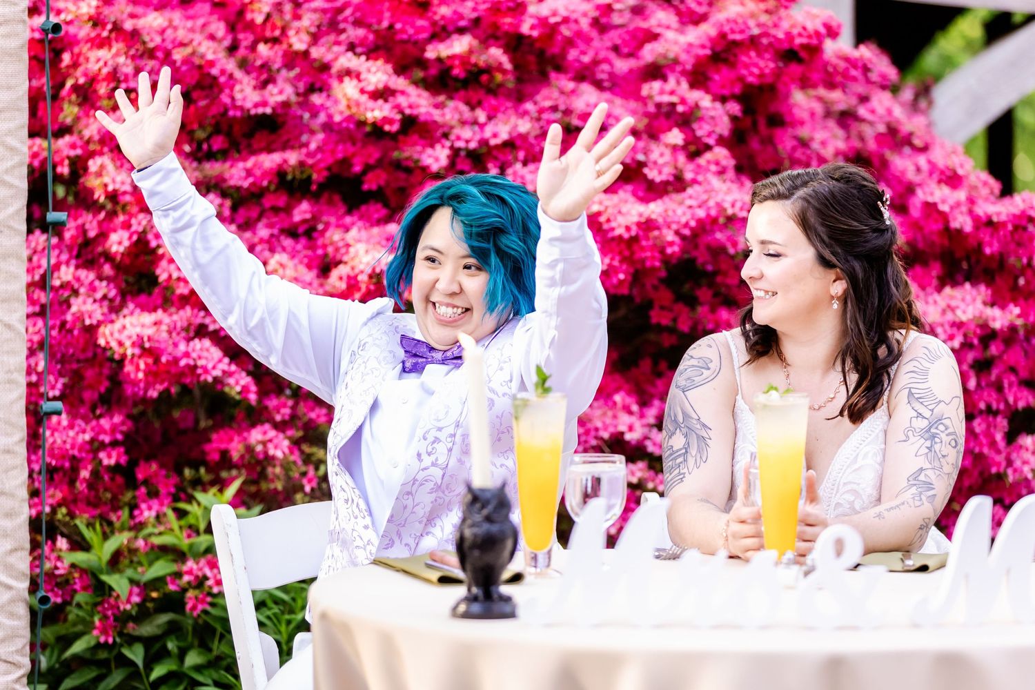 A celebratory outdoor party scene with pink azalea bushes and yellow drinks on a white tablecloth.