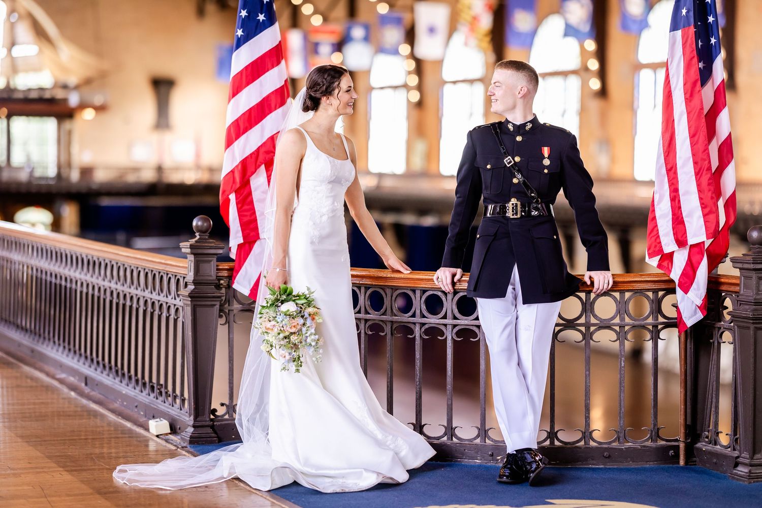 A couple in formal attire stands between American flags in an elegant indoor venue.