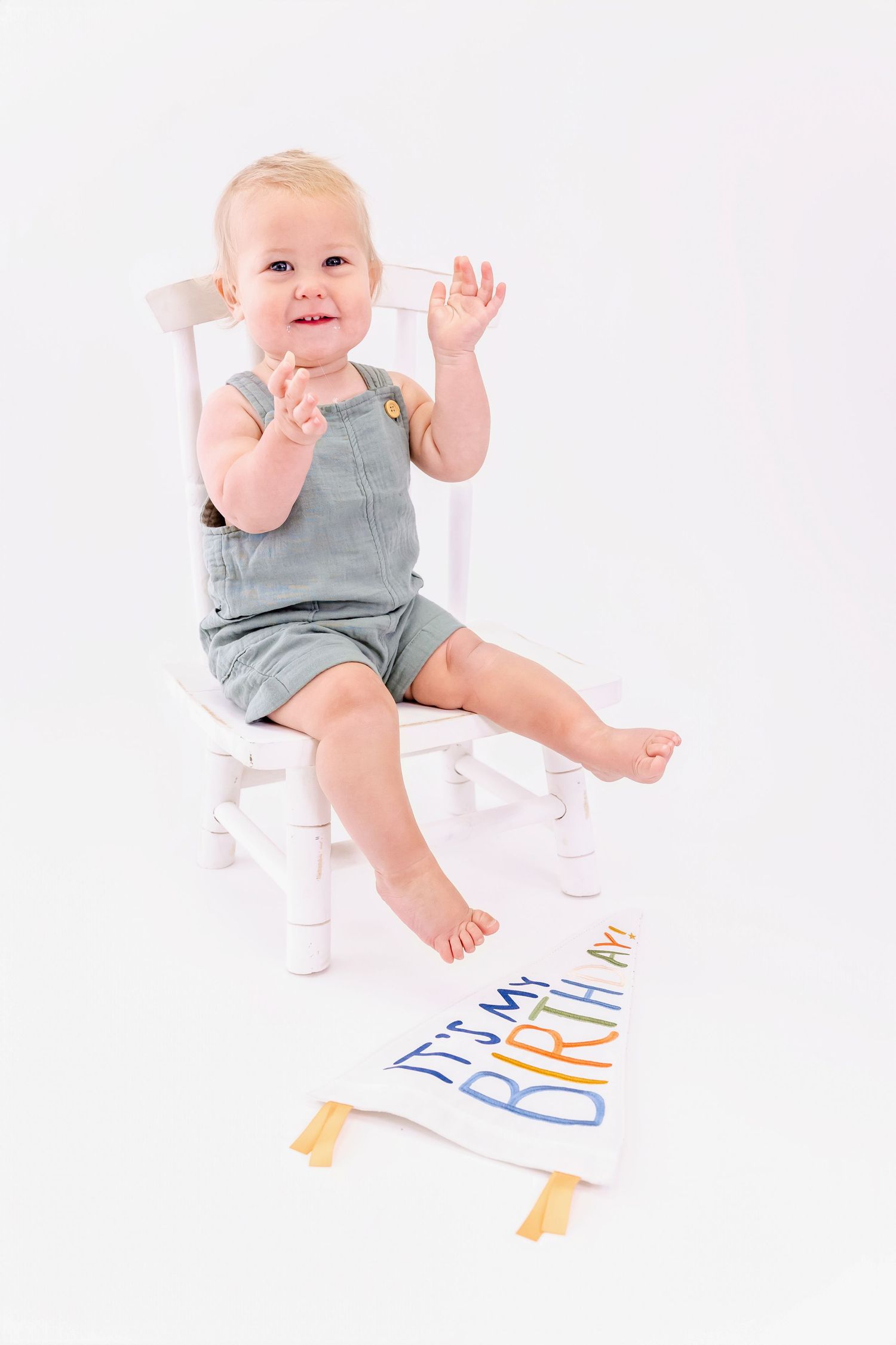 A baby in grey overalls sits on a white chair with birthday decorations below.