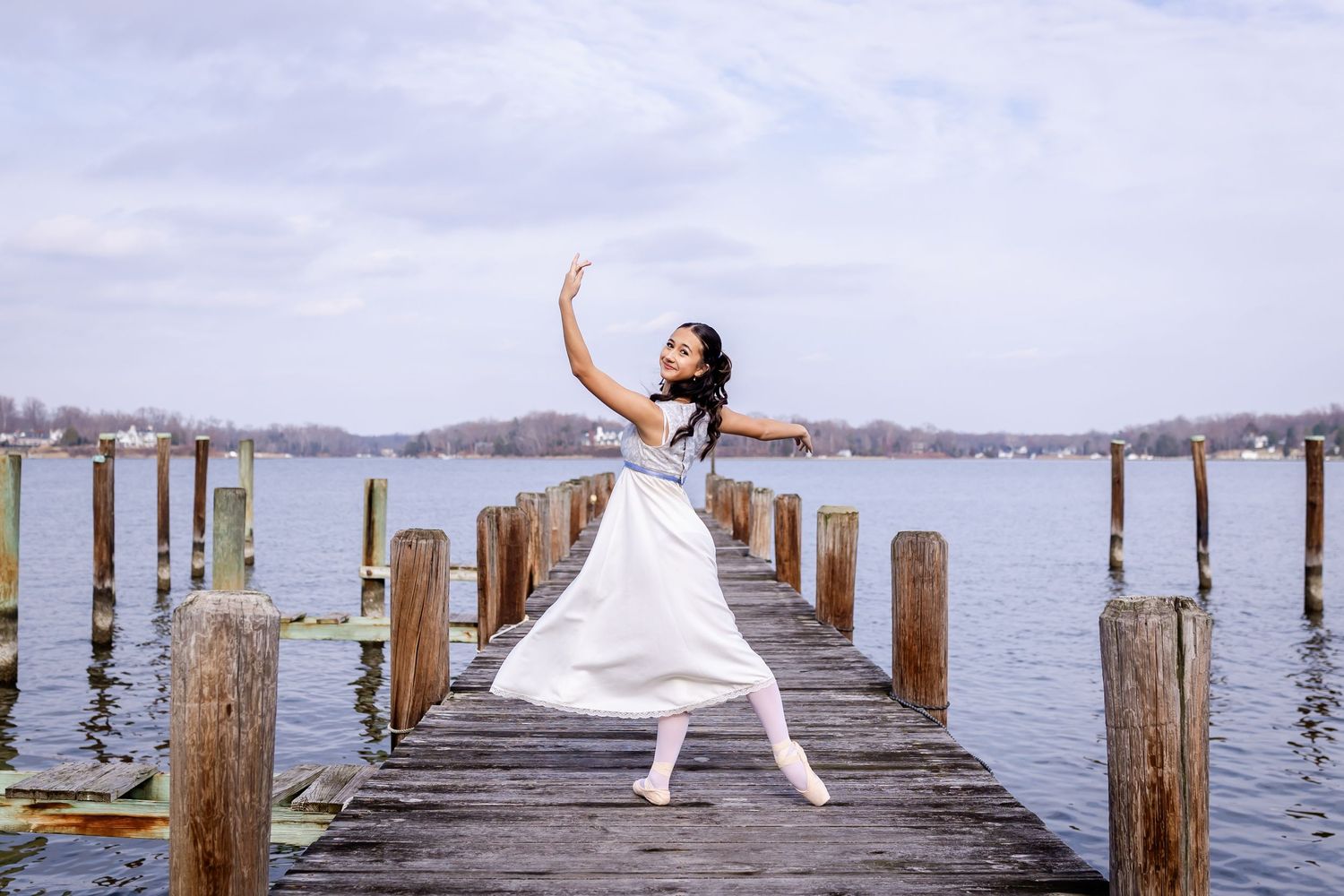 Dancer in white performing graceful poses on a wooden dock by the water.