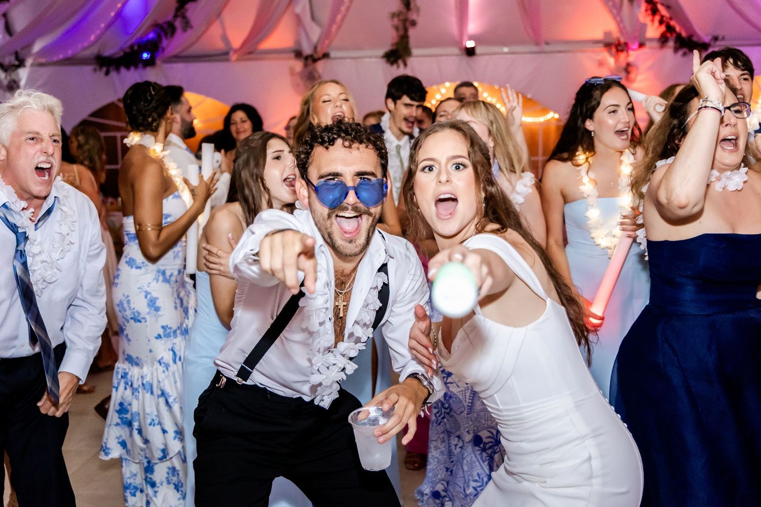 Wedding guests dance and celebrate on a crowded dance floor with colorful purple lighting at a reception party.