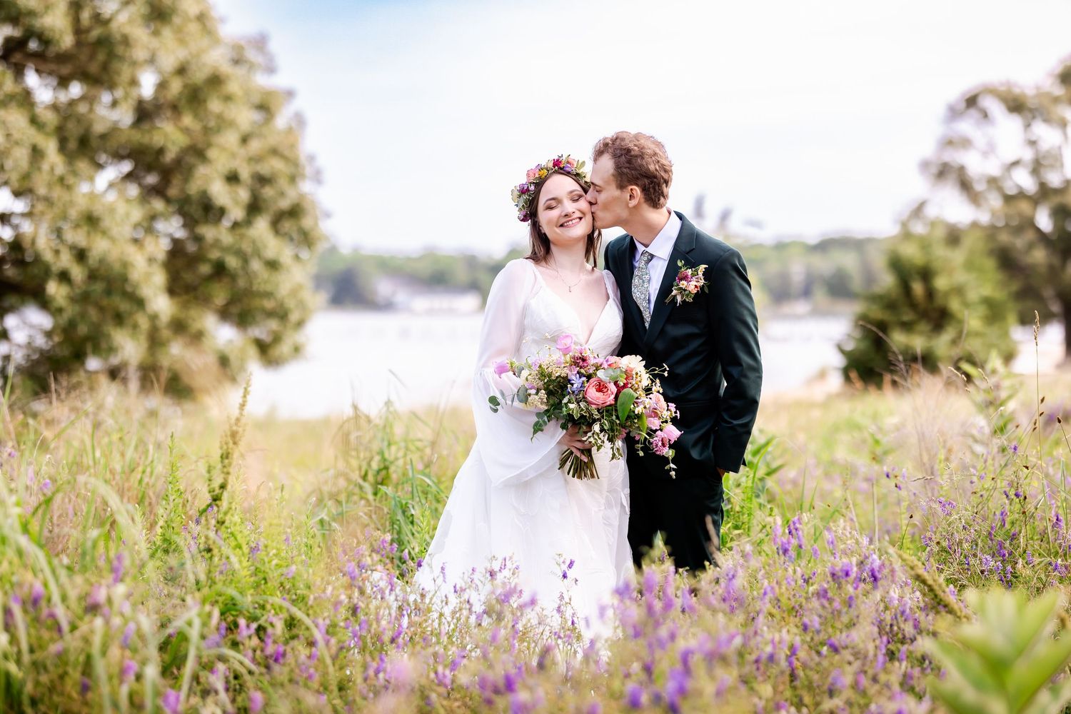 A couple shares a romantic moment among purple wildflowers by a serene lakeside on their wedding day.