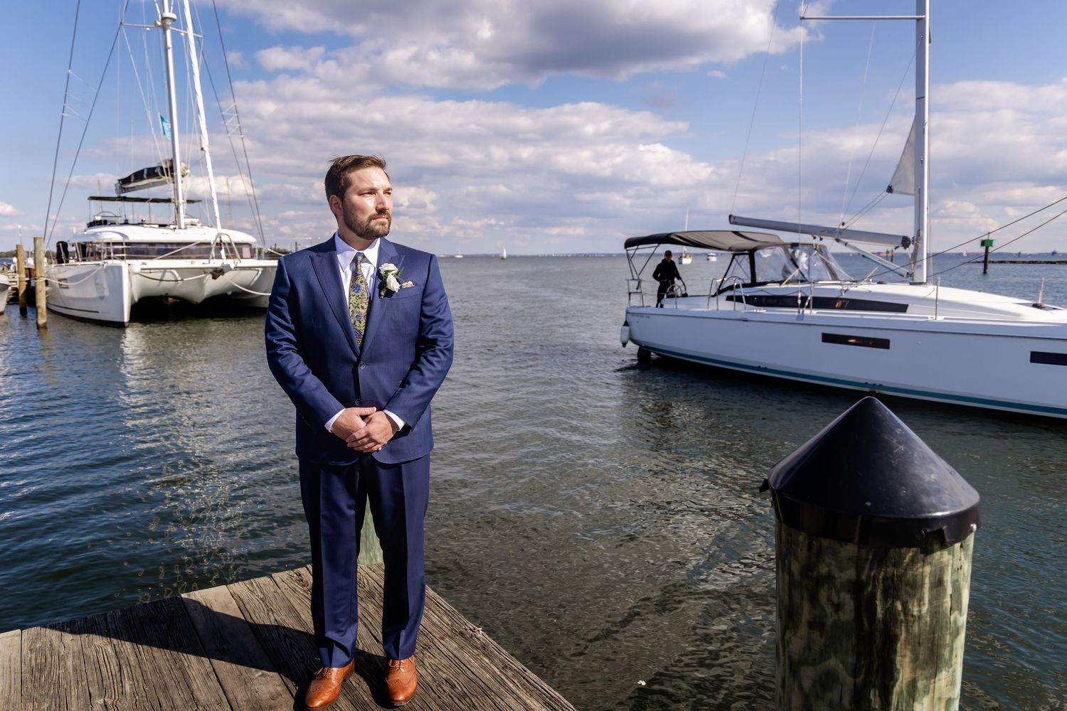 Elegant man in blue suit standing on wooden dock with sailboats moored in calm harbor waters behind him.