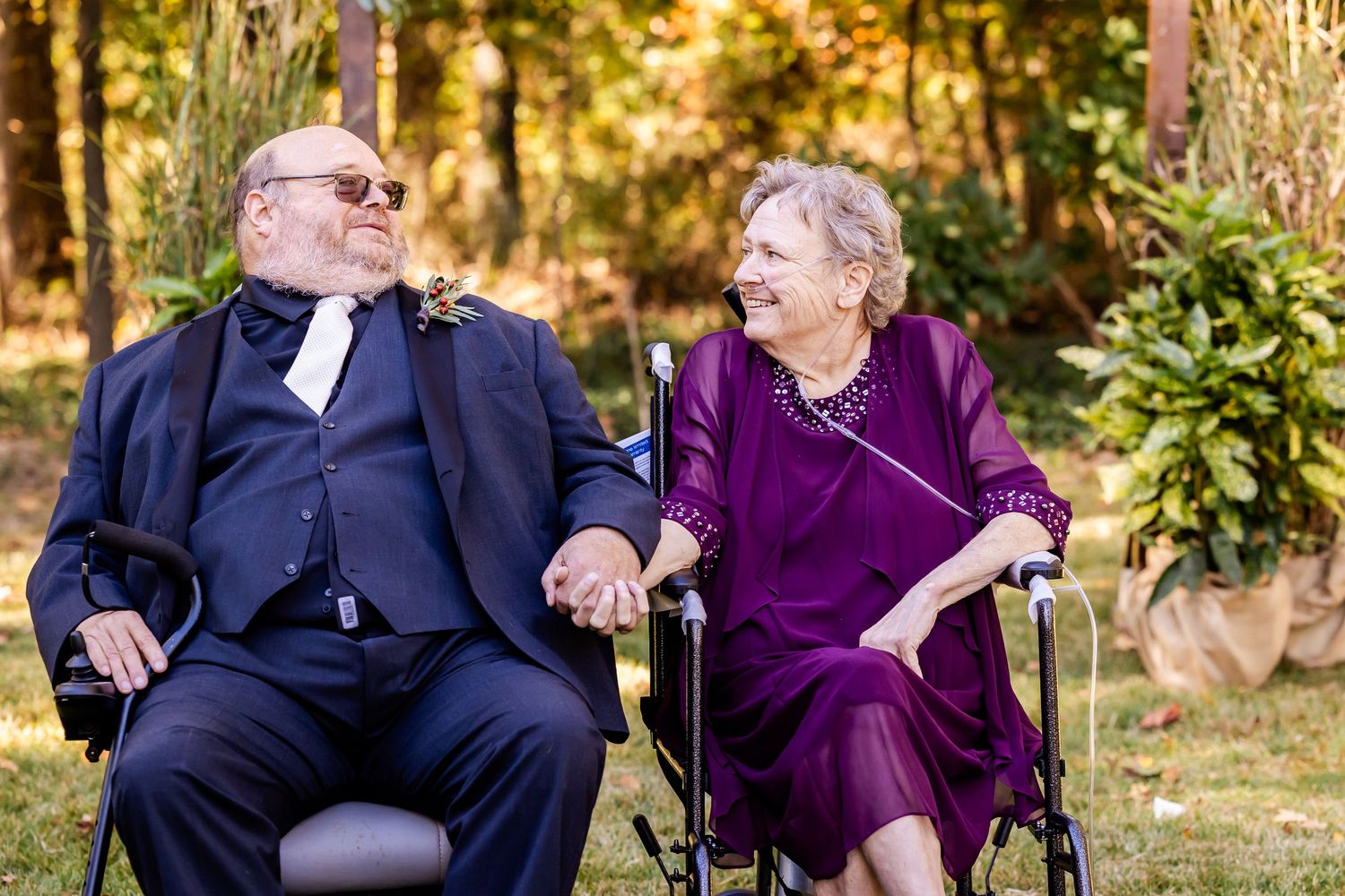 An elderly couple in formal attire share a tender moment while sitting together outdoors.