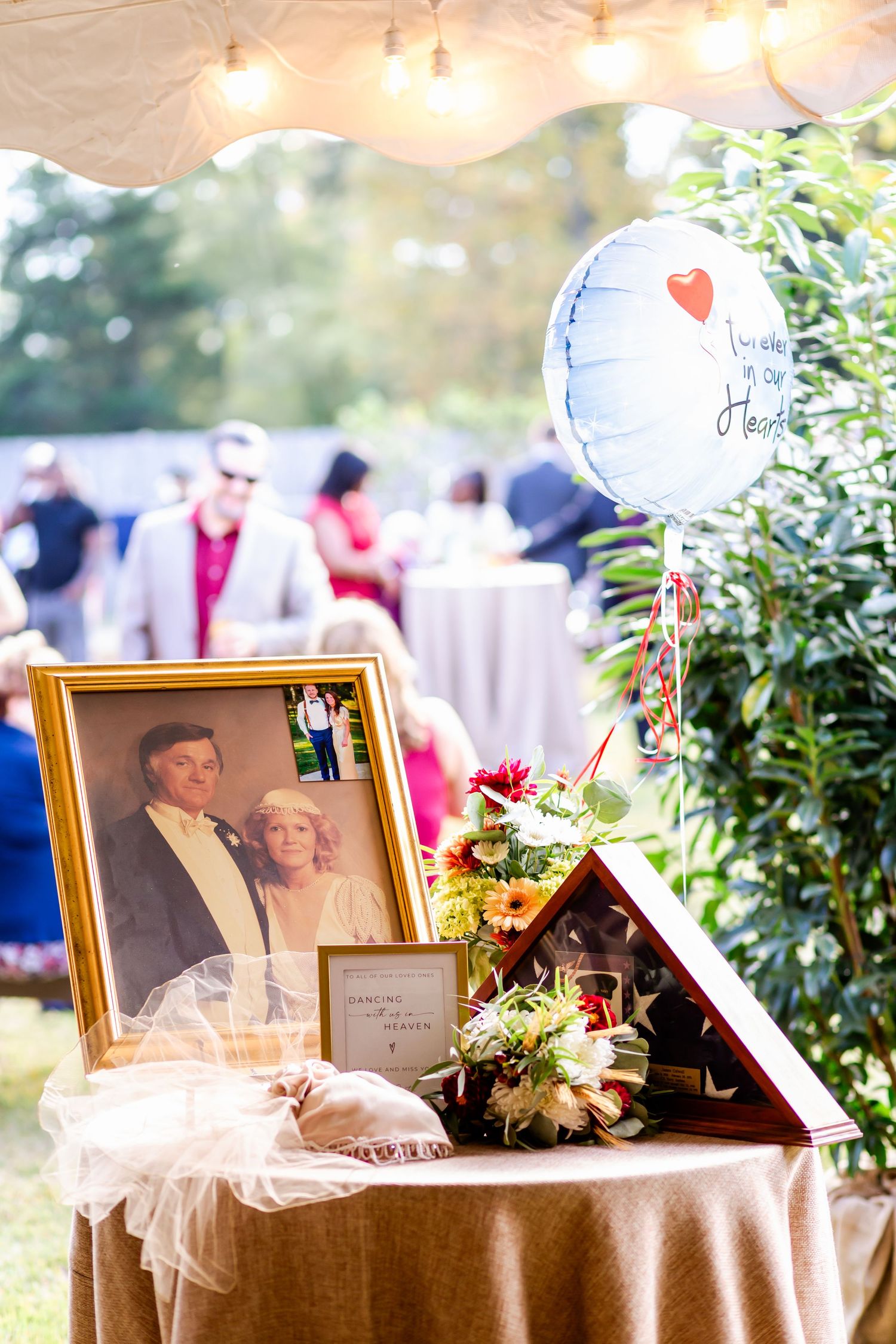 Memorial display with framed photographs and floral arrangements on a table at an outdoor event.